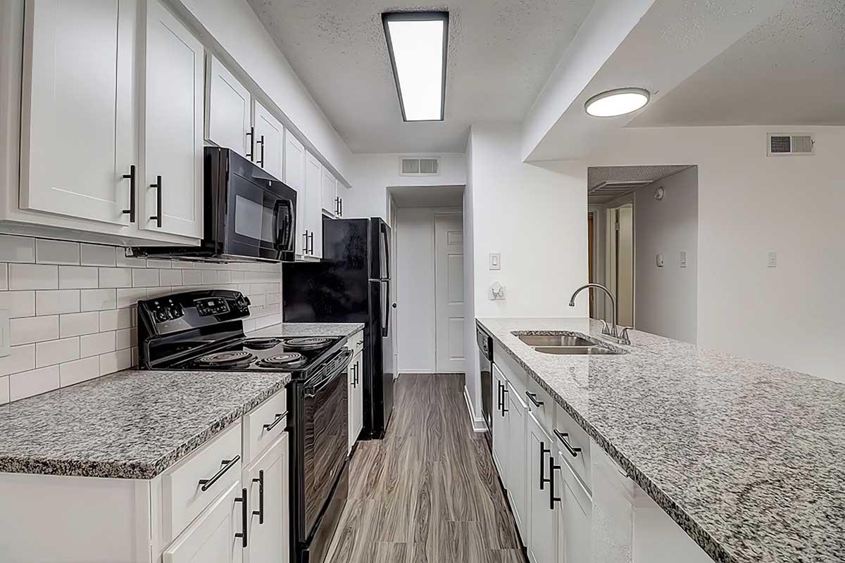 A modern kitchen with white cabinets, black appliances, and a granite countertop. There is a stove, oven, microwave, and sink, along with a doorway leading to another room. The floor features a wood-like finish, and the overall design is sleek and contemporary, emphasizing open space and light.
