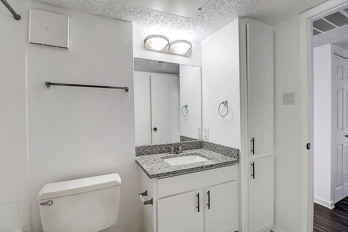 A modern bathroom featuring a white toilet, a granite countertop with a sink, and a large mirror above it. The walls are white, and there is a towel rack and a door leading to a separate area. The lighting is bright, enhancing the clean and minimalist design.