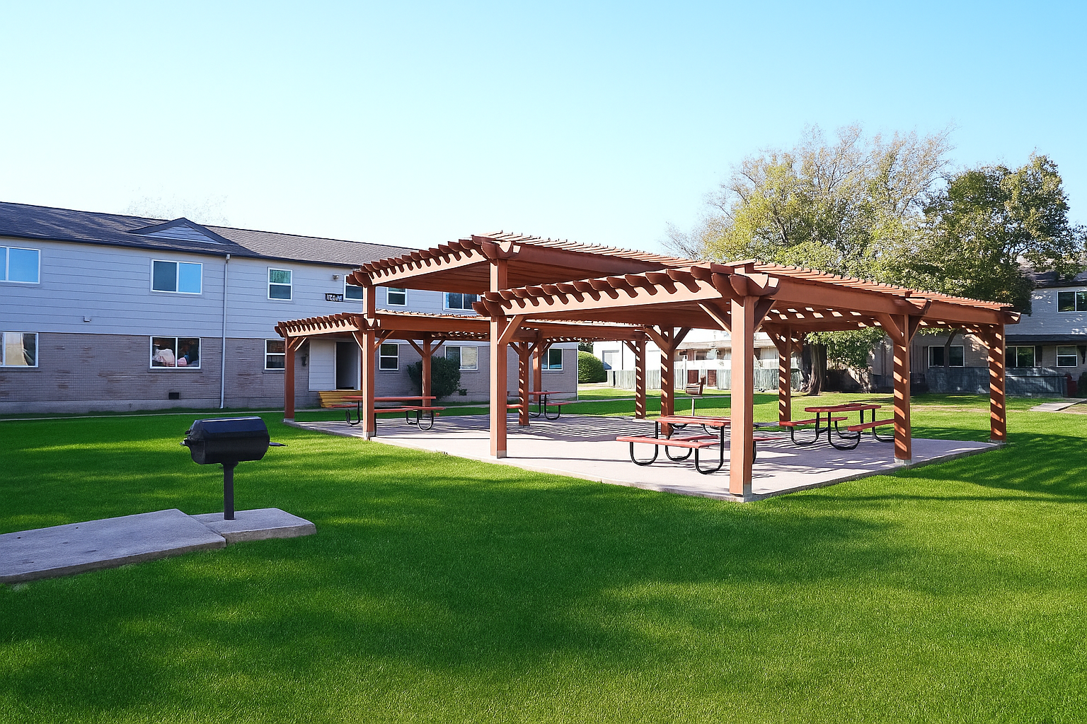 A sunny residential area featuring two wooden pergolas with picnic tables underneath, set on a green lawn. Nearby, there's a charcoal grill. The backdrop includes a multi-story building with windows and trees in the distance, creating a serene outdoor space.