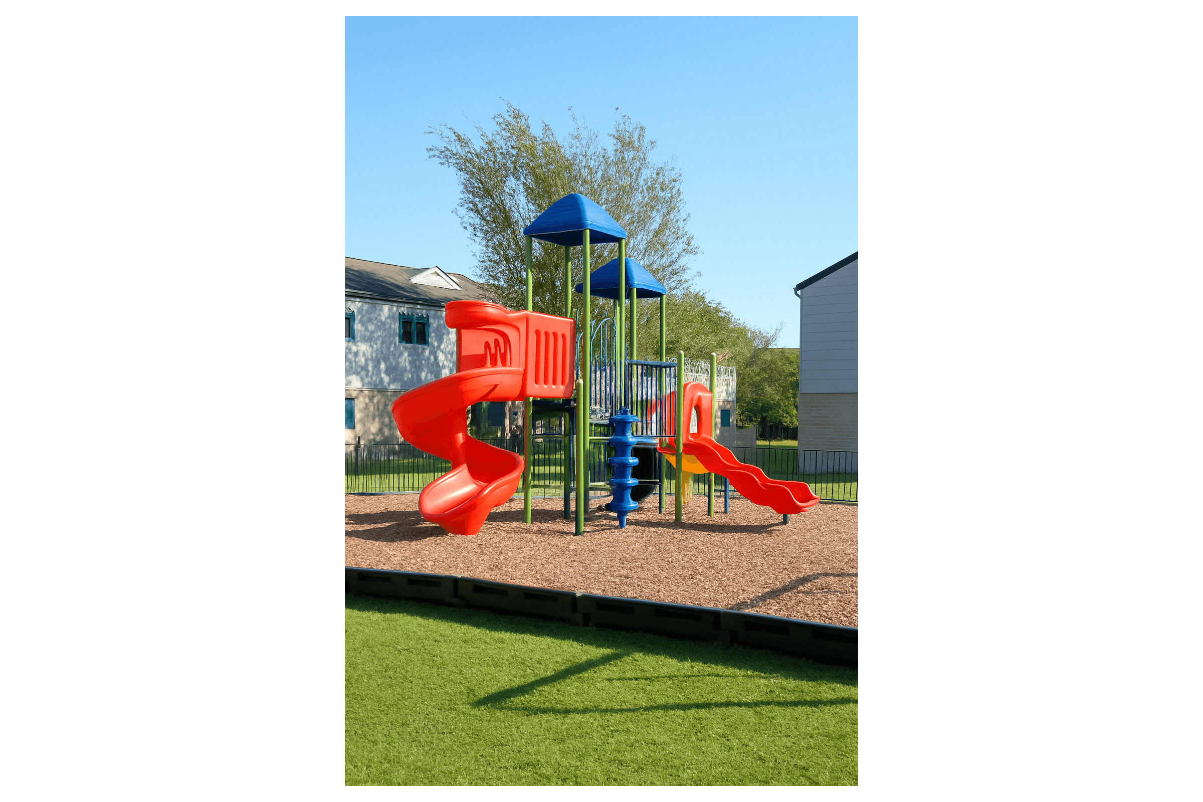 Colorful playground equipment featuring a red slide, climbing structures, and blue elements, set on a grassy area with a gravel surface. The playground is surrounded by trees and residential buildings, under a clear blue sky.