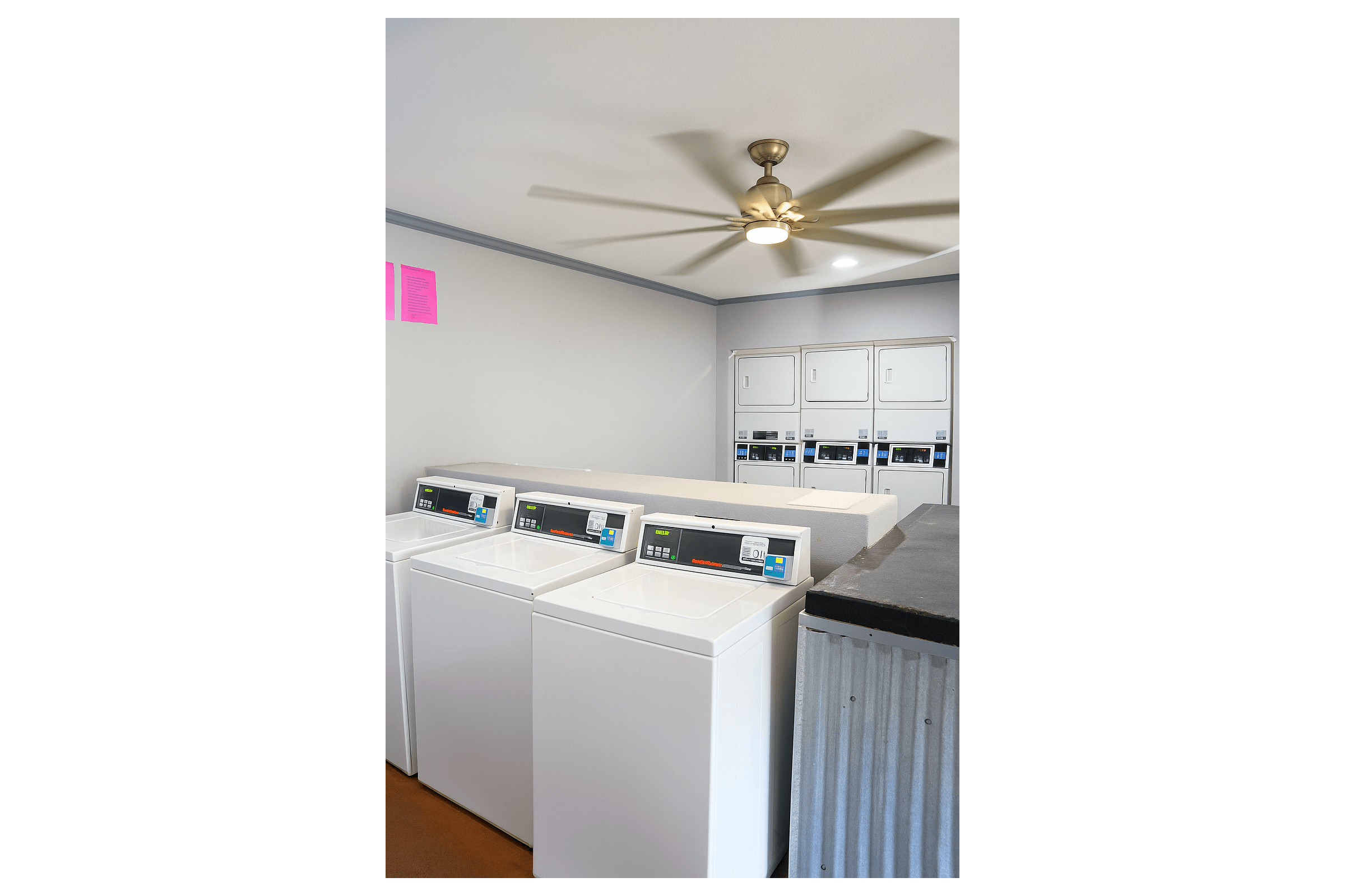 A clean laundry room featuring several white washing machines and a row of dryer units. The ceiling has a fan, and the walls are light gray. There are informational signs in pink on the wall. The environment is well-lit and organized, providing a functional space for laundry tasks.