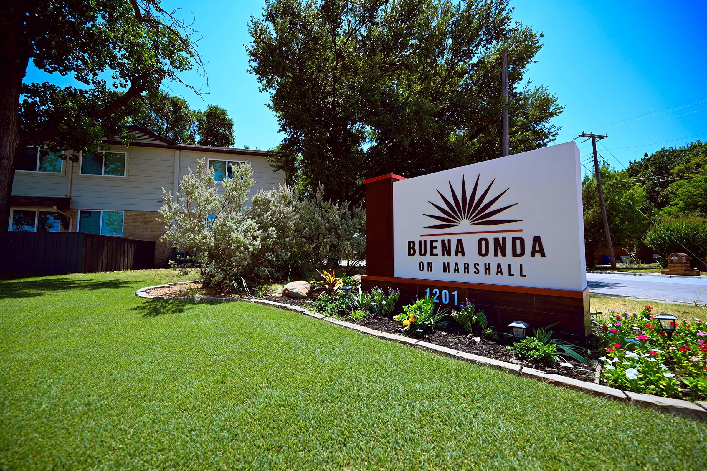 A sign for "Buena Onda on Marshall" is prominently displayed in front of a landscaped area with plants and flowers. The sign features a sunburst design and the address "1201." The setting includes green grass and trees in the background, suggesting a welcoming environment.