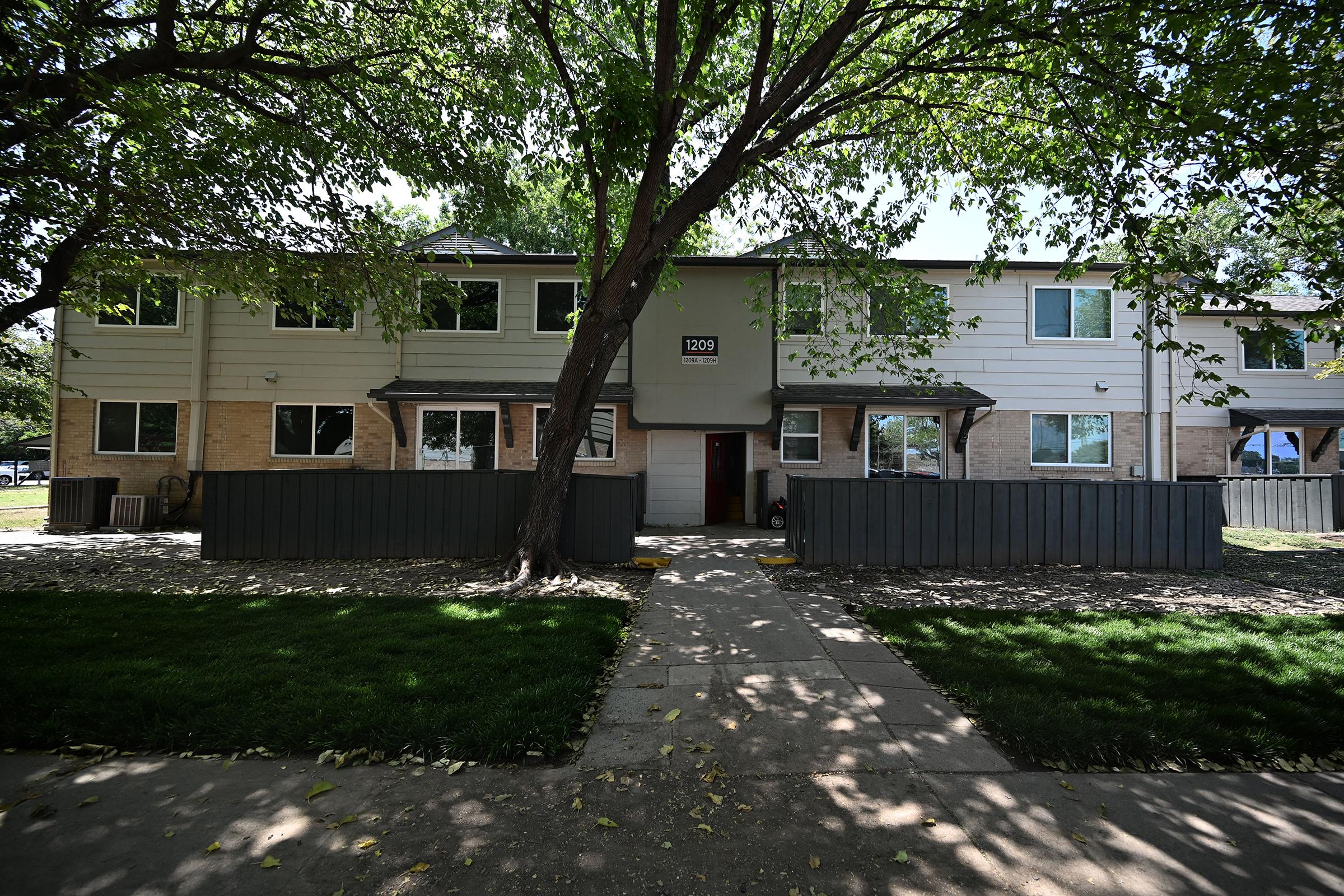 A two-story residential building with multiple windows, surrounded by trees and a grassy area. A pathway leads to the entrance, with a wooden fence in the foreground. The building features a neutral color scheme and is well-lit by natural sunlight.