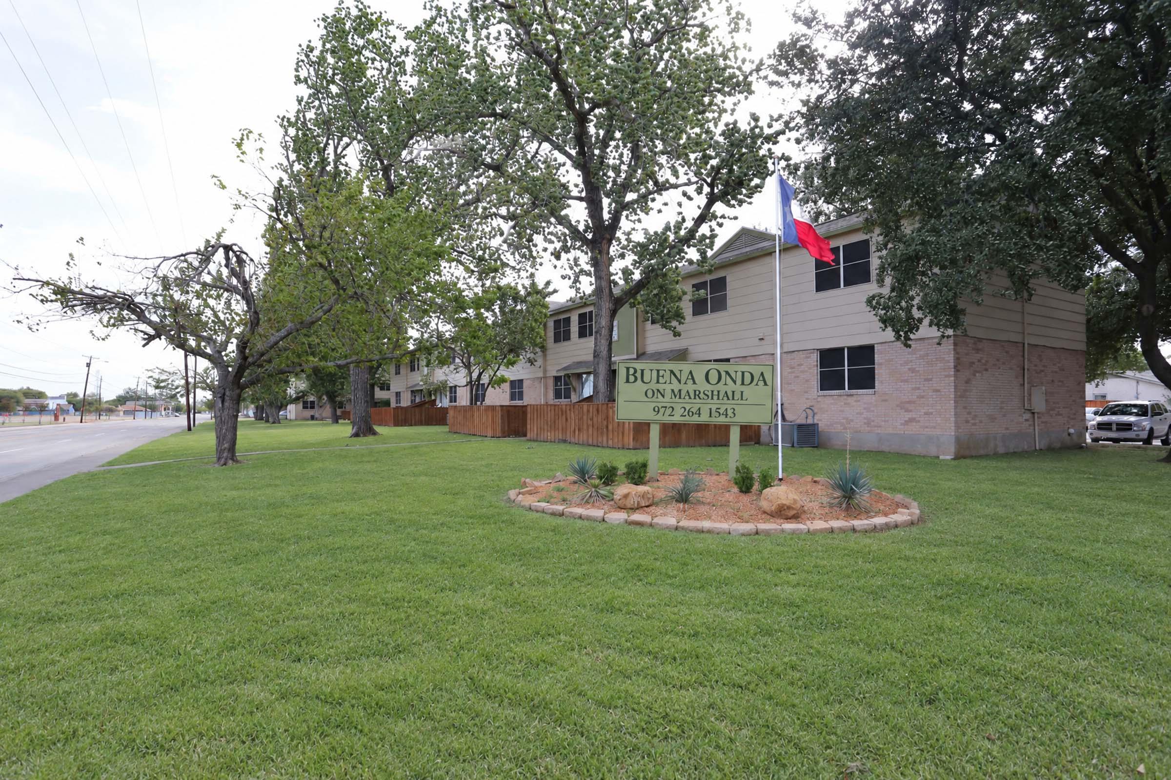 A view of a residential building with a sign saying "Buena Onda on Marshall." The building is surrounded by well-maintained grass and trees. A Texas flag is prominently displayed. The setting appears to be an urban or suburban area with a road nearby.