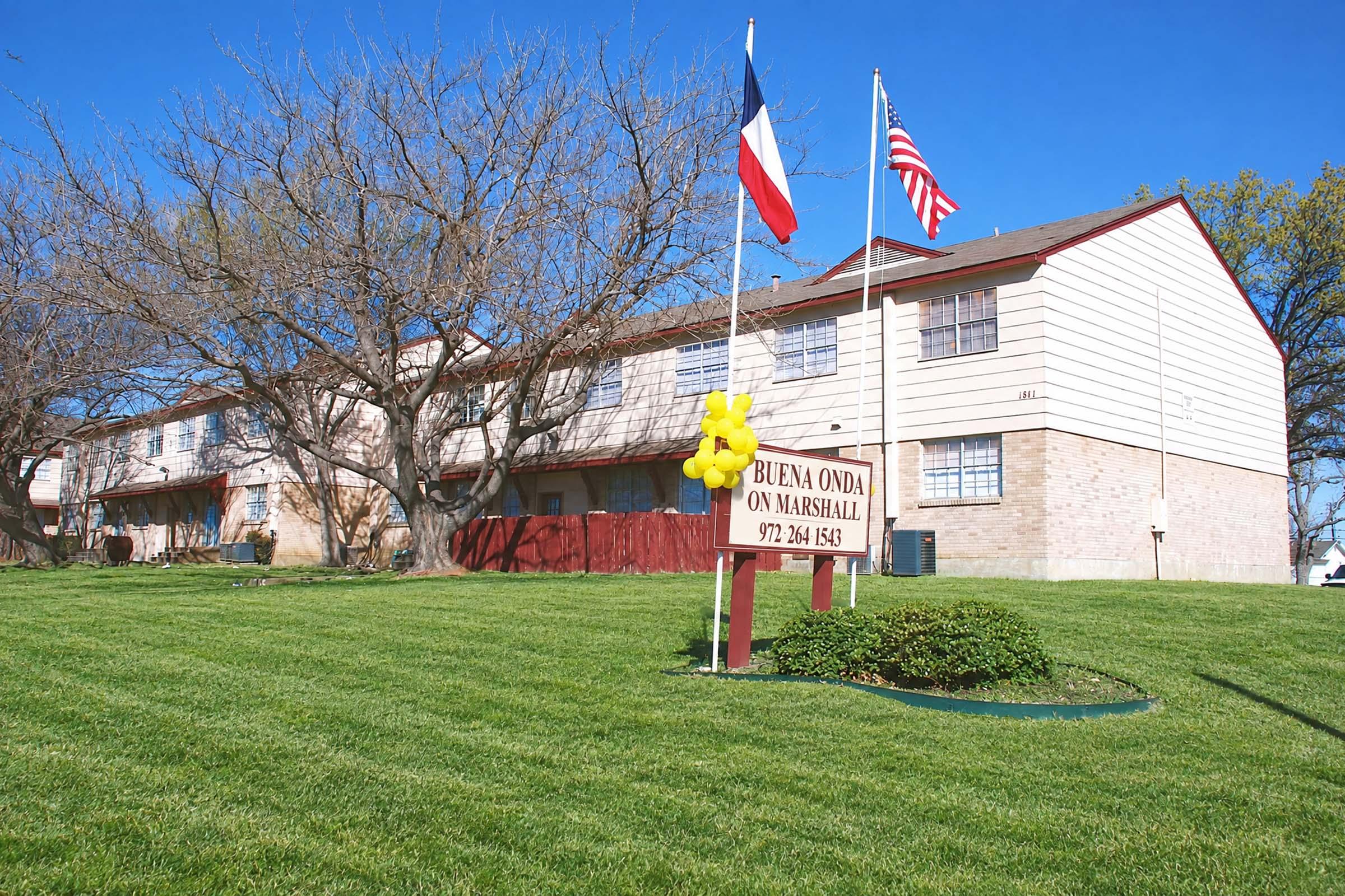 A two-story residential building with a well-maintained lawn. The exterior features a tan and white color scheme. Flags of Texas and the United States are displayed on poles in front of the building, along with a sign that reads “Buena Onda on Marshall” with contact information. Surrounding trees are bare, indicating early spring or winter.