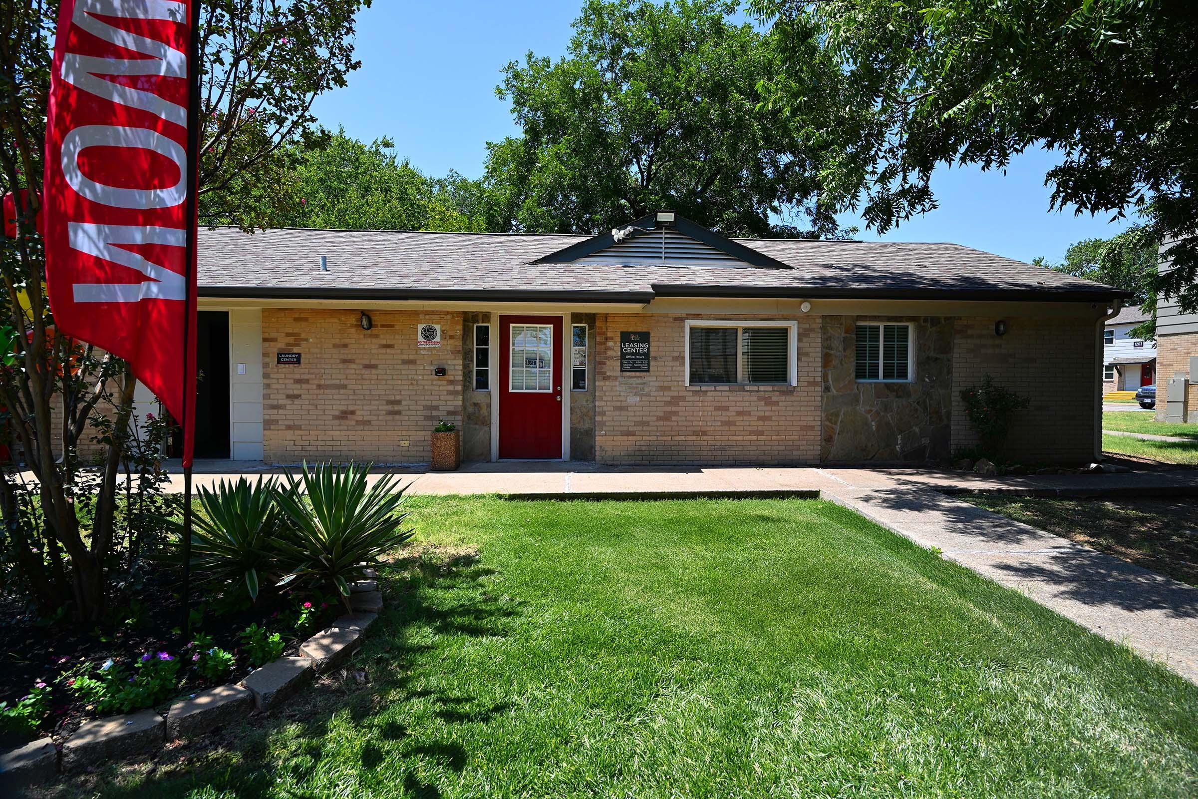 Single-story brick house with a red door, surrounded by green grass and shrubs. A large "NOW" flag is displayed prominently in front. The home features several windows and a well-maintained yard, providing a welcoming and bright appearance.