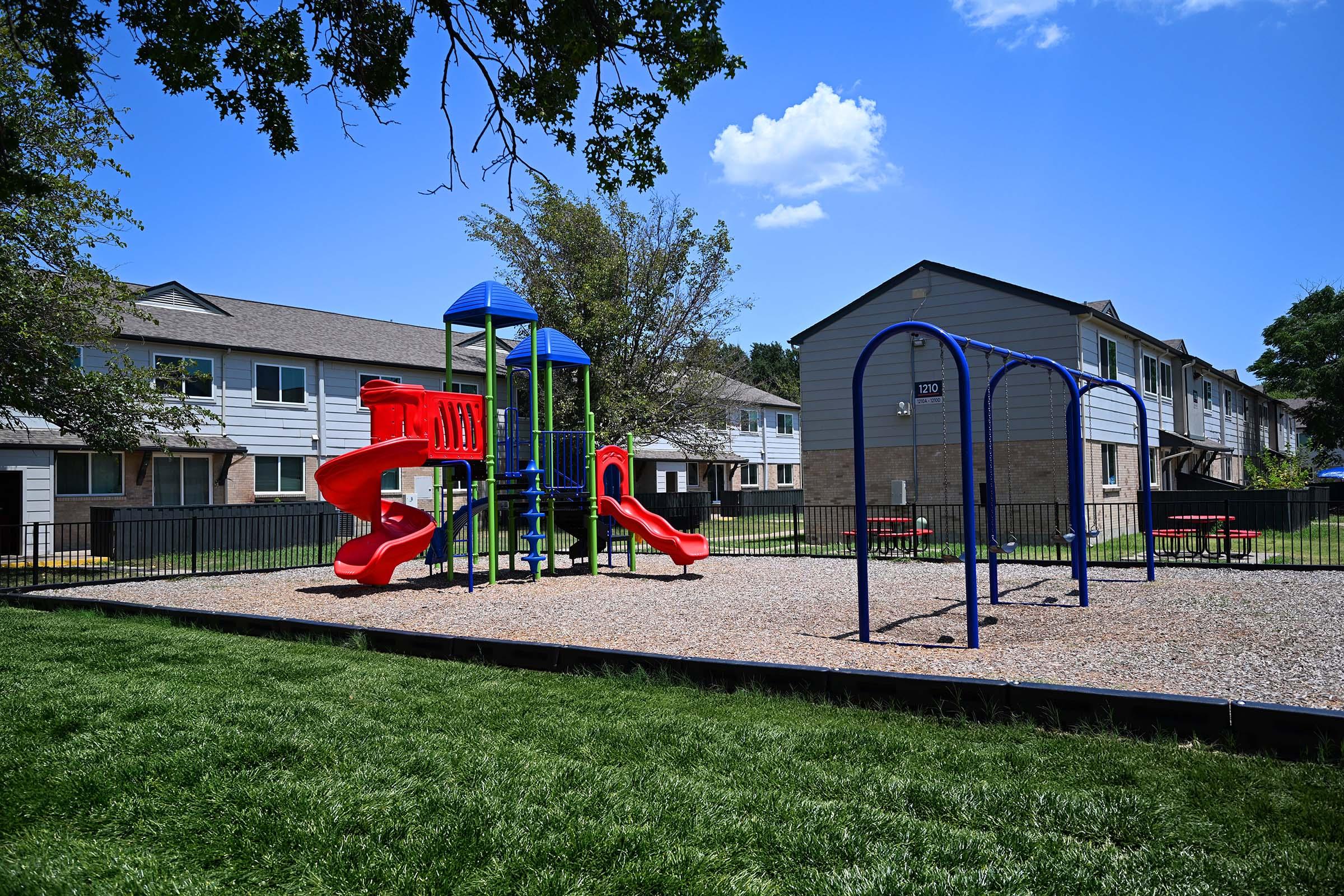 A colorful playground with a red slide, climbing structure, and blue swings, situated on a grassy area. In the background, there are several residential buildings with light-colored exteriors and trees. The sky is clear with a few clouds.
