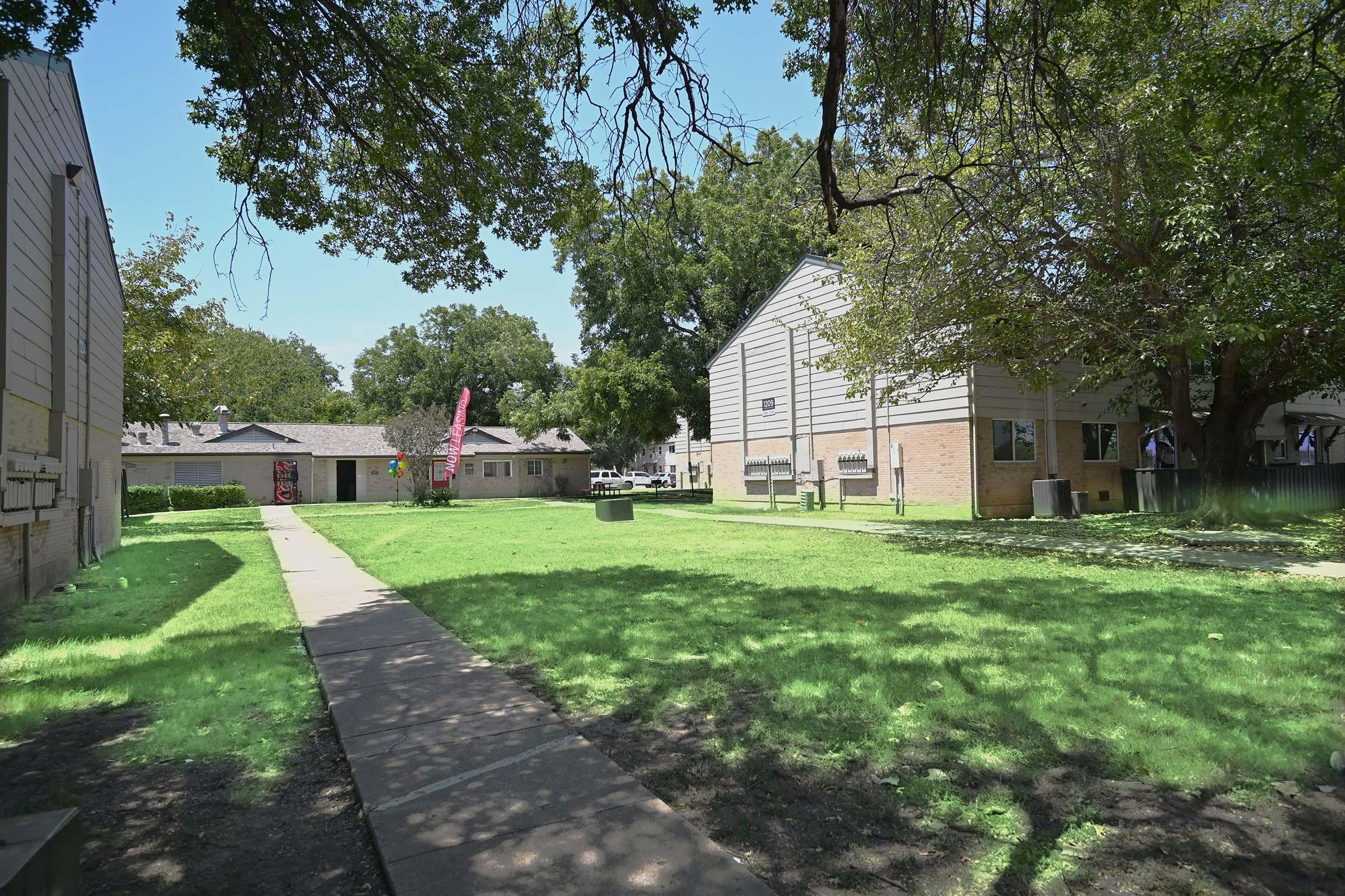 A sunny outdoor scene showing a grassy area between two multi-story residential buildings. The pathway runs through the lawn, surrounded by trees. There are benches and a vibrant pink flag visible in the background, adding to the inviting atmosphere.