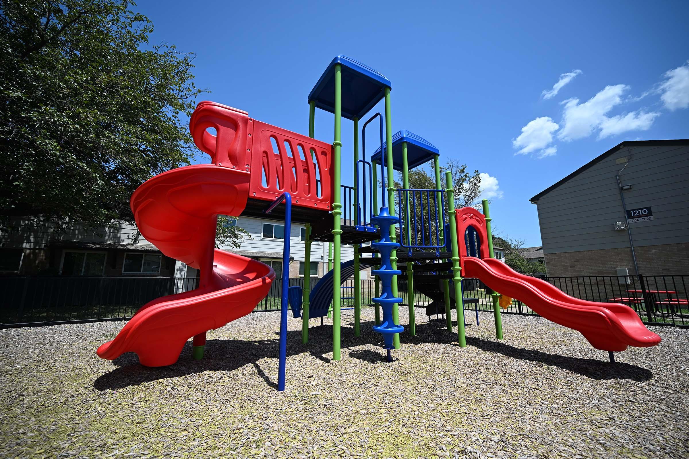 A colorful playground structure featuring red and blue slides, climbing equipment, and a lookout tower, set in a gravel area with green trees in the background under a clear blue sky. Ideal for children to play and explore.