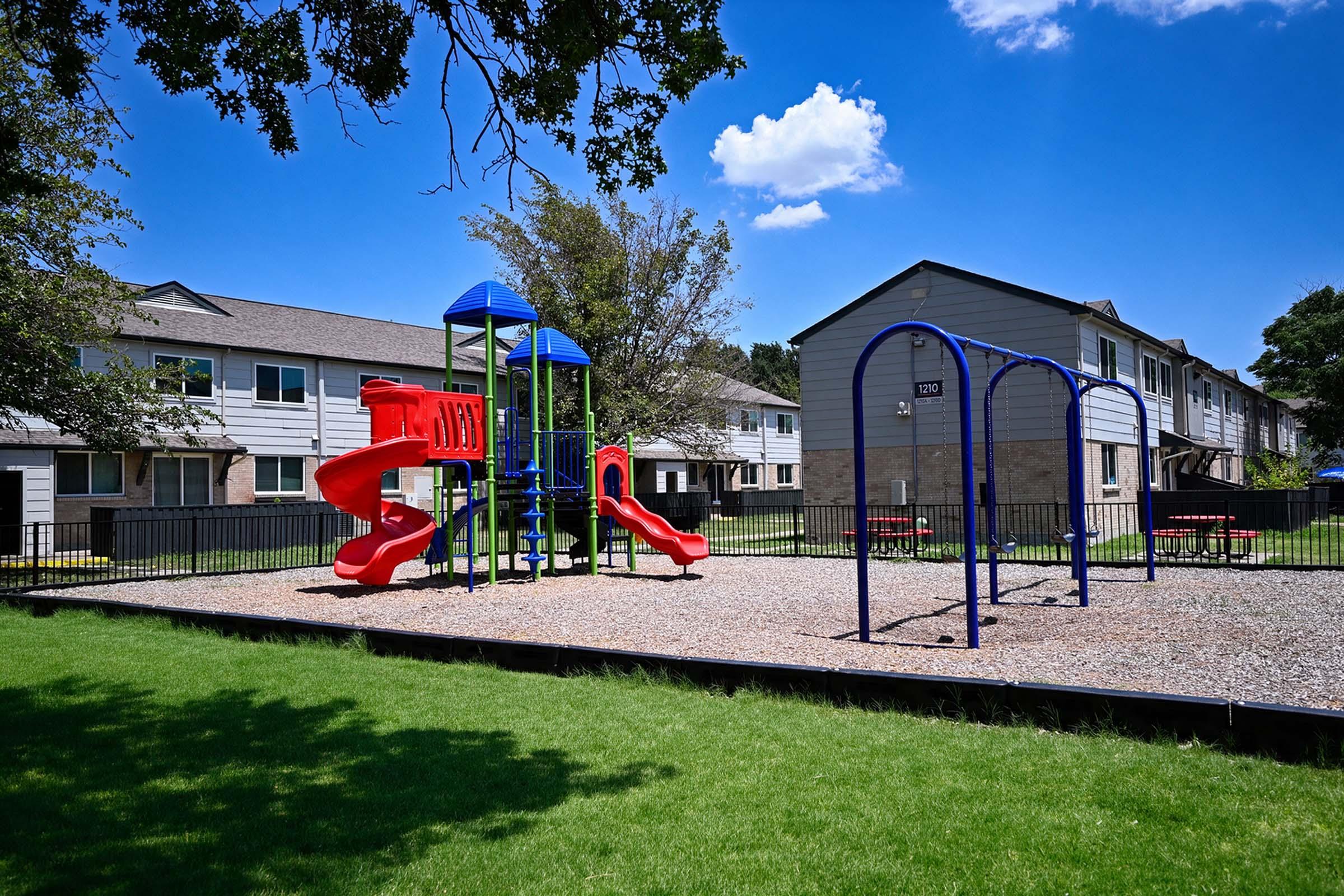 A colorful playground featuring red slides and climbing structures, accompanied by blue swings. The playground is surrounded by a grassy area and is situated behind several residential buildings under a clear blue sky with a few clouds.