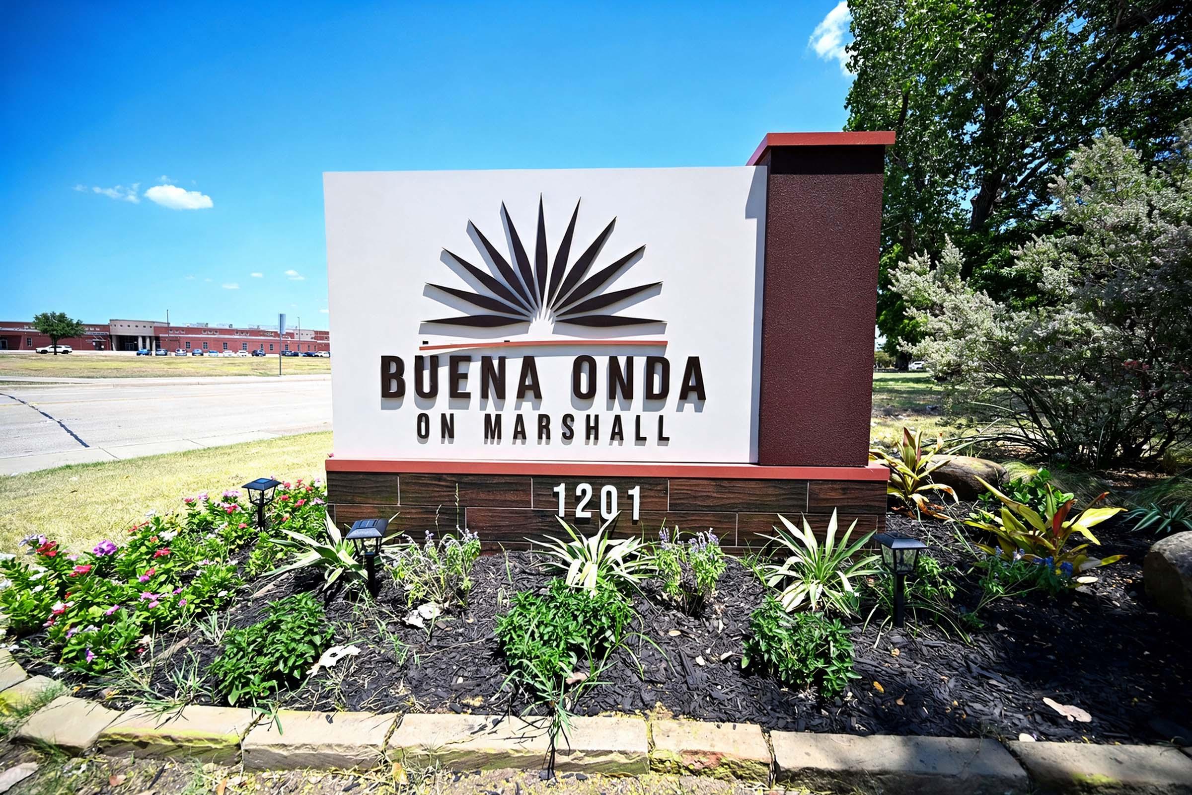 Sign for "Buena Onda on Marshall," featuring stylized sunburst design, located at 1201 Marshall. Surrounded by colorful landscaping, including plants and flowers, with a clear blue sky in the background.