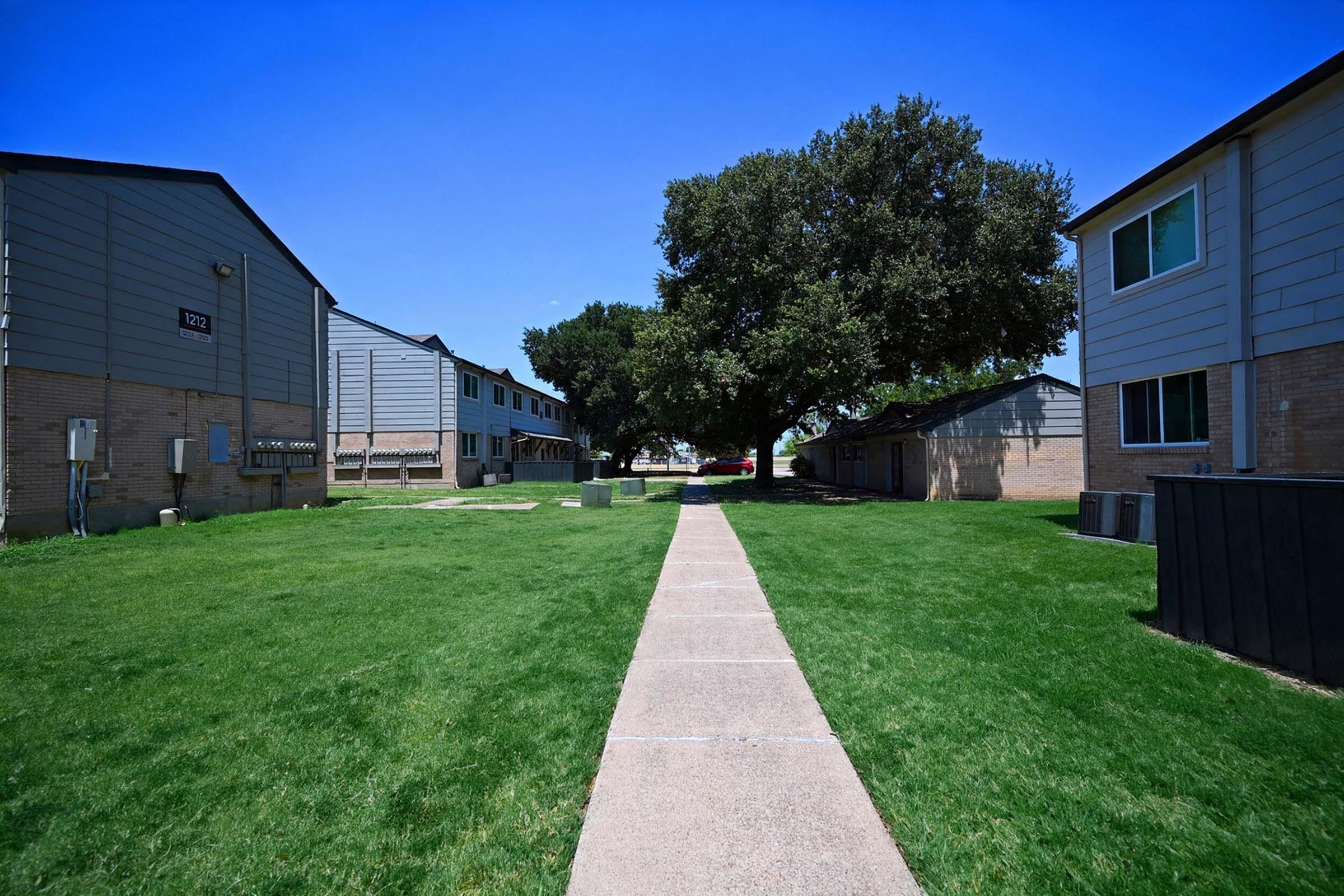 A clear, sunny day shows a walkway lined by grassy areas between residential buildings. On one side, two-story apartment units are visible with a large tree providing shade. The scene conveys a peaceful, suburban atmosphere with a blue sky overhead.