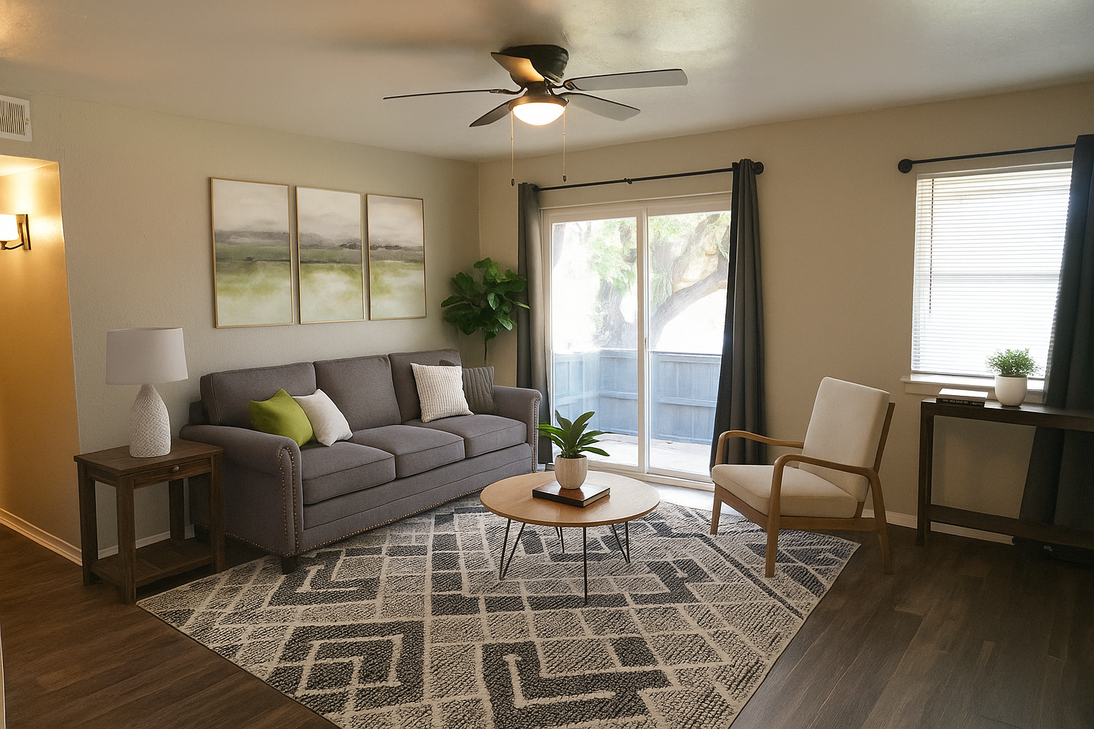 A cozy living room featuring a gray sofa with green and white accent pillows, a round wooden coffee table, and a comfortable armchair. Natural light fills the space through a sliding glass door leading to a patio. There is a large potted plant, a small table with a decorative item, and artwork on the wall.