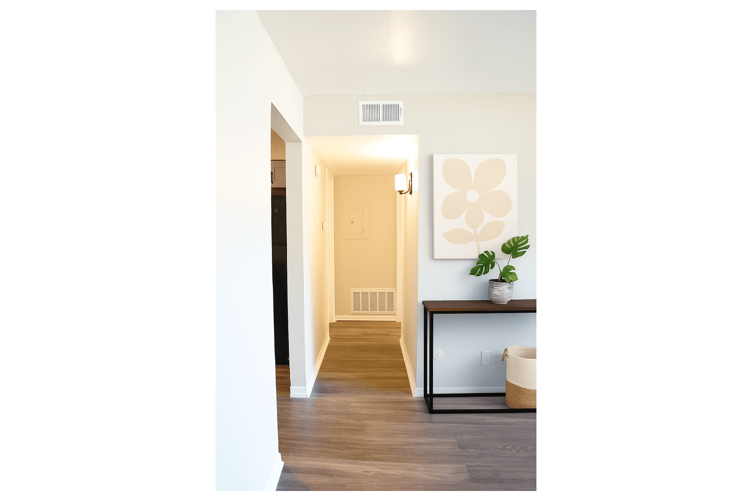 A brightly lit hallway featuring wooden flooring, a light-colored wall, and modern decor. On the left, there is a chair and a table with a plant, while the right side displays a large artwork with a floral design. The space appears inviting and well-maintained.