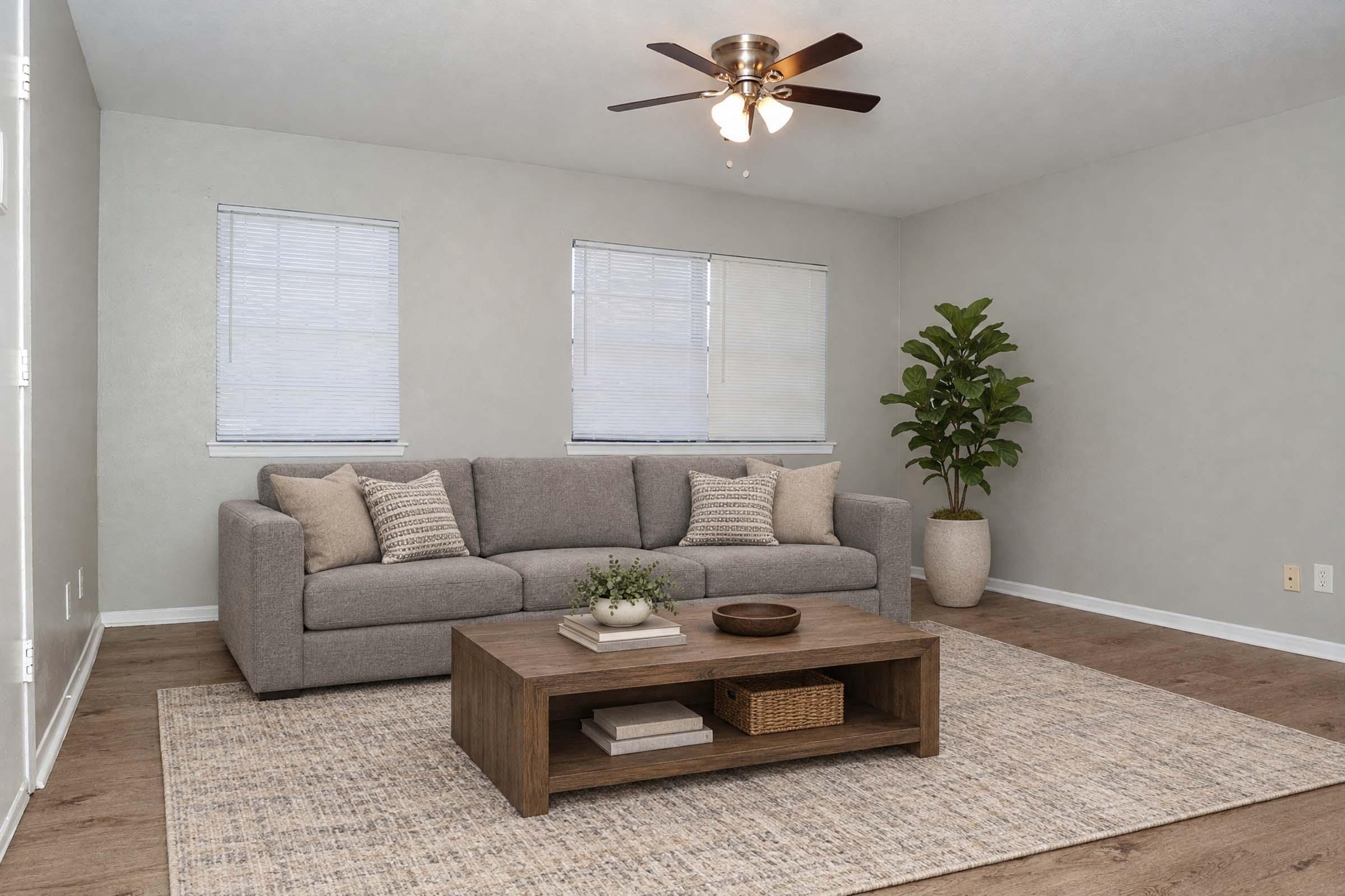 A modern living room featuring a gray sofa with decorative pillows, a wooden coffee table with a few accessories, and a indoor plant in the corner. There are two windows with blinds allowing natural light to enter, and the floor is covered with a light area rug, all set against neutral gray walls.
