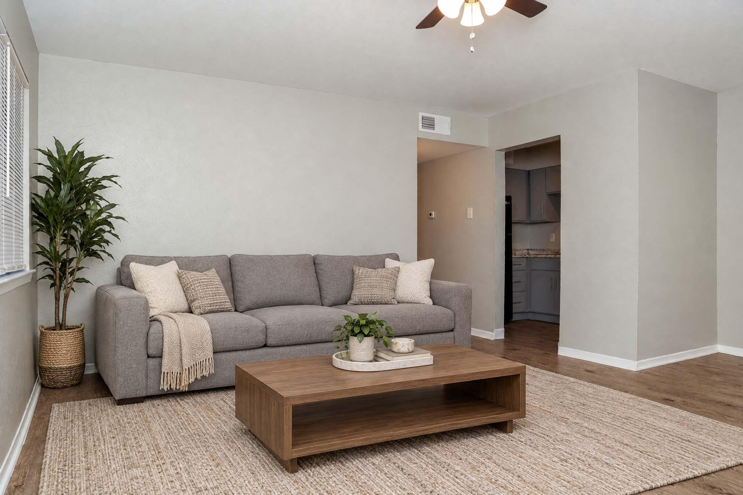 A cozy living room featuring a gray sofa adorned with pillows, a wooden coffee table with a plant on it, and a light-colored area rug. There's a potted plant in the corner and a view into a kitchen in the background. The walls are painted a soft gray, and a ceiling fan adds to the inviting atmosphere.