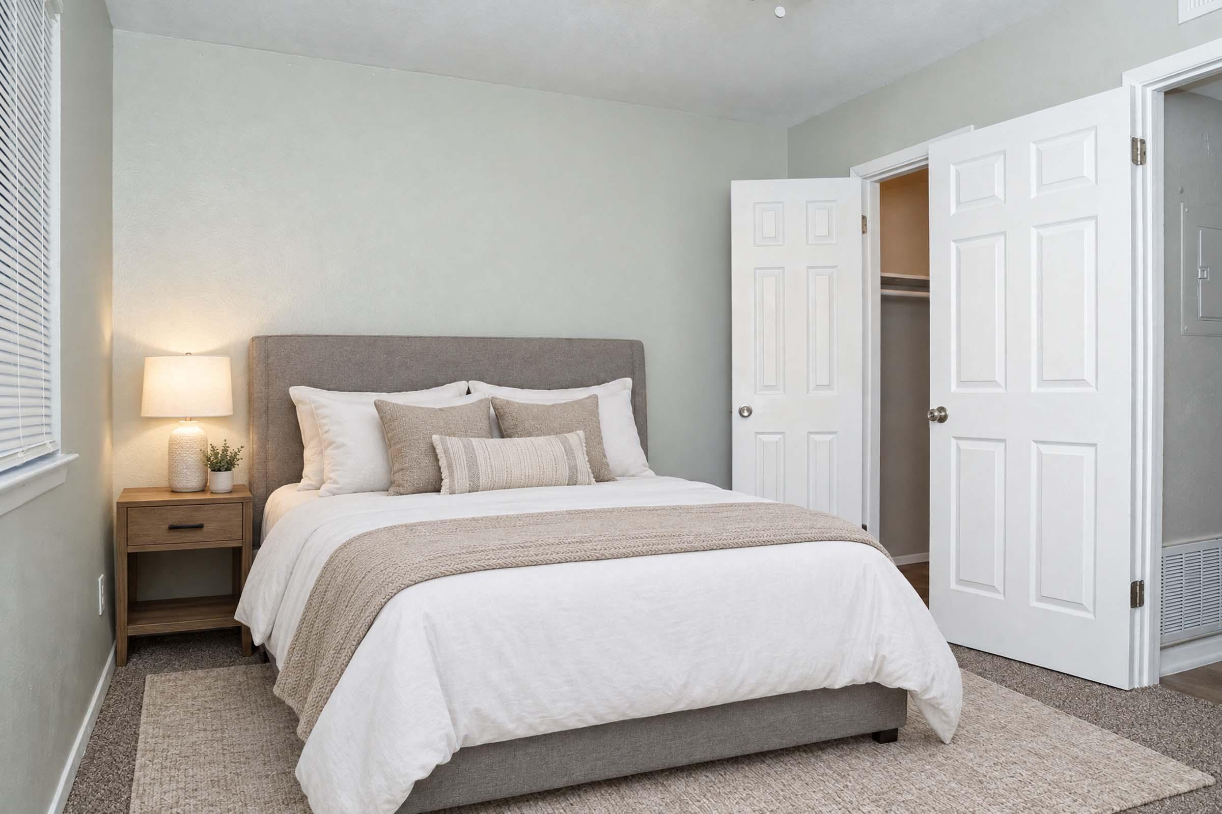 A cozy bedroom featuring a queen-sized bed with white bedding and a taupe throw blanket. A bedside table with a small plant and a lamp is next to the bed. There are light green walls, a window with blinds, and a closet door partially open on the right. The floor is covered with a soft carpet.