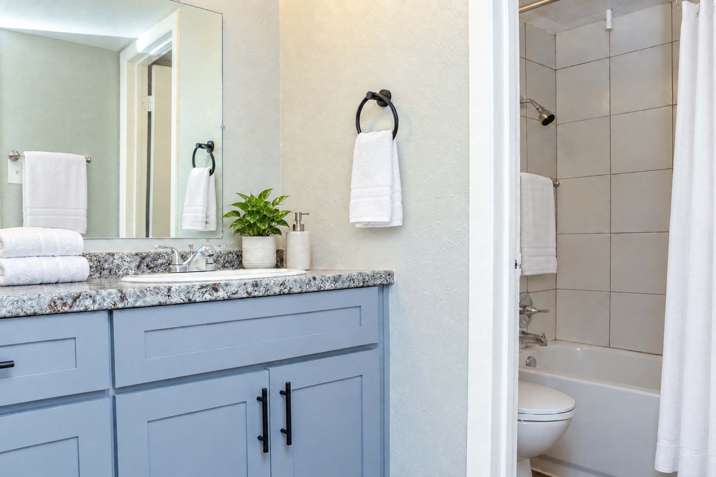 Modern bathroom featuring a granite countertop with a sink, towels hung on a rack, a small plant, and a shower area. The color scheme includes soft gray cabinetry and white tiles, with a clean and organized appearance.