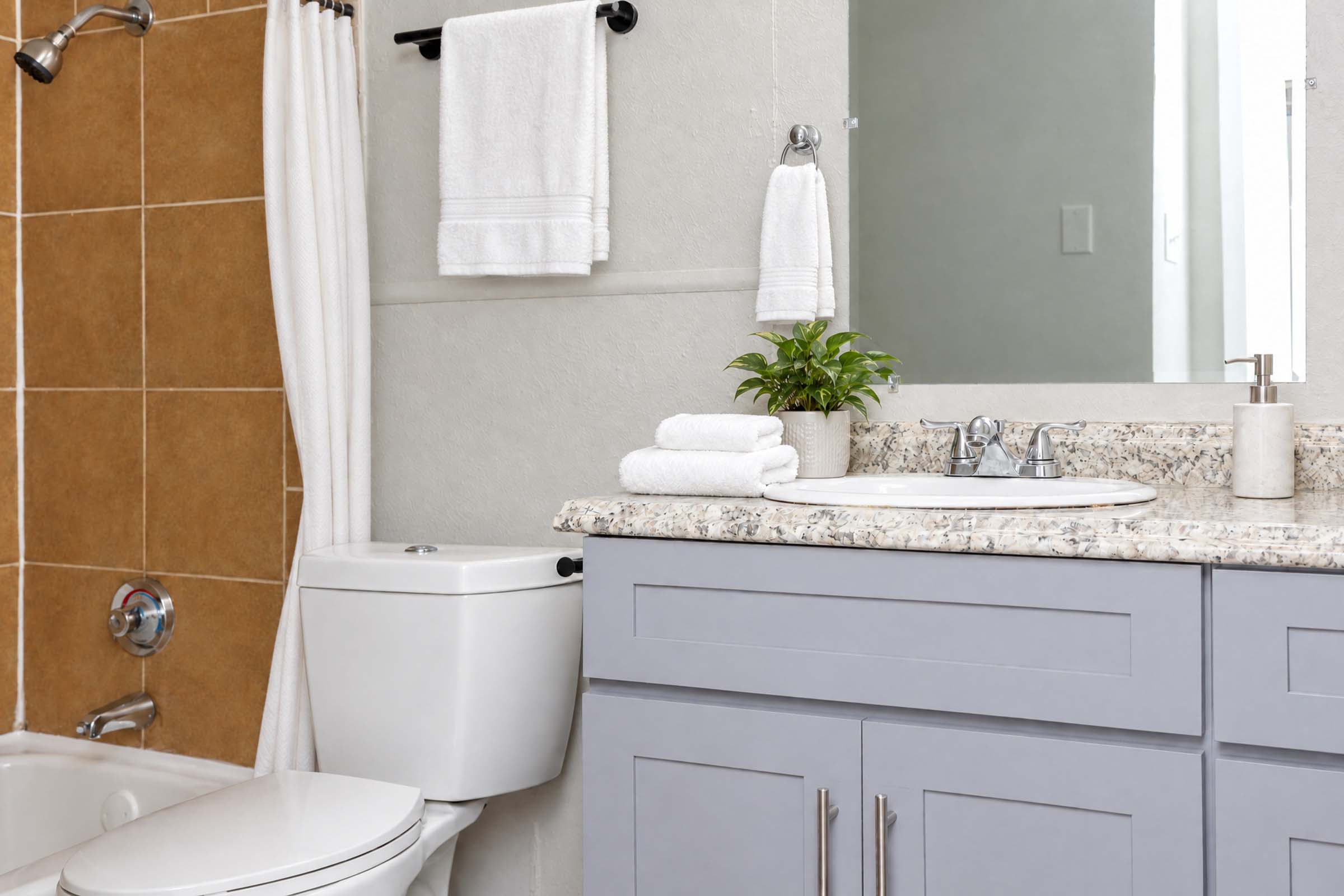 A modern bathroom featuring a light gray vanity with a marble countertop, neatly arranged white towels, and a small green plant. The wall has beige tiles, and there is a white toilet beside the vanity. A mirror reflects light, enhancing the airy atmosphere of the space.