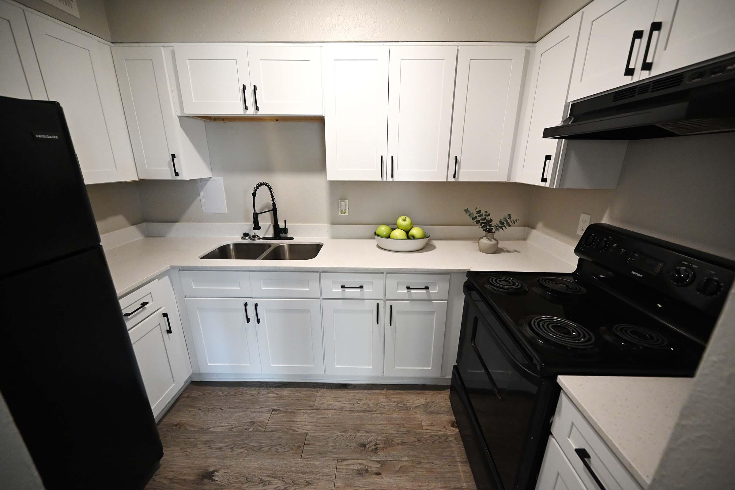 A modern kitchen featuring white cabinets, a black refrigerator, a black stove, and a black sink. The countertop is light-colored with a bowl of green apples and a small plant as decor. The floor is made of light wood, contributing to a bright and clean aesthetic.