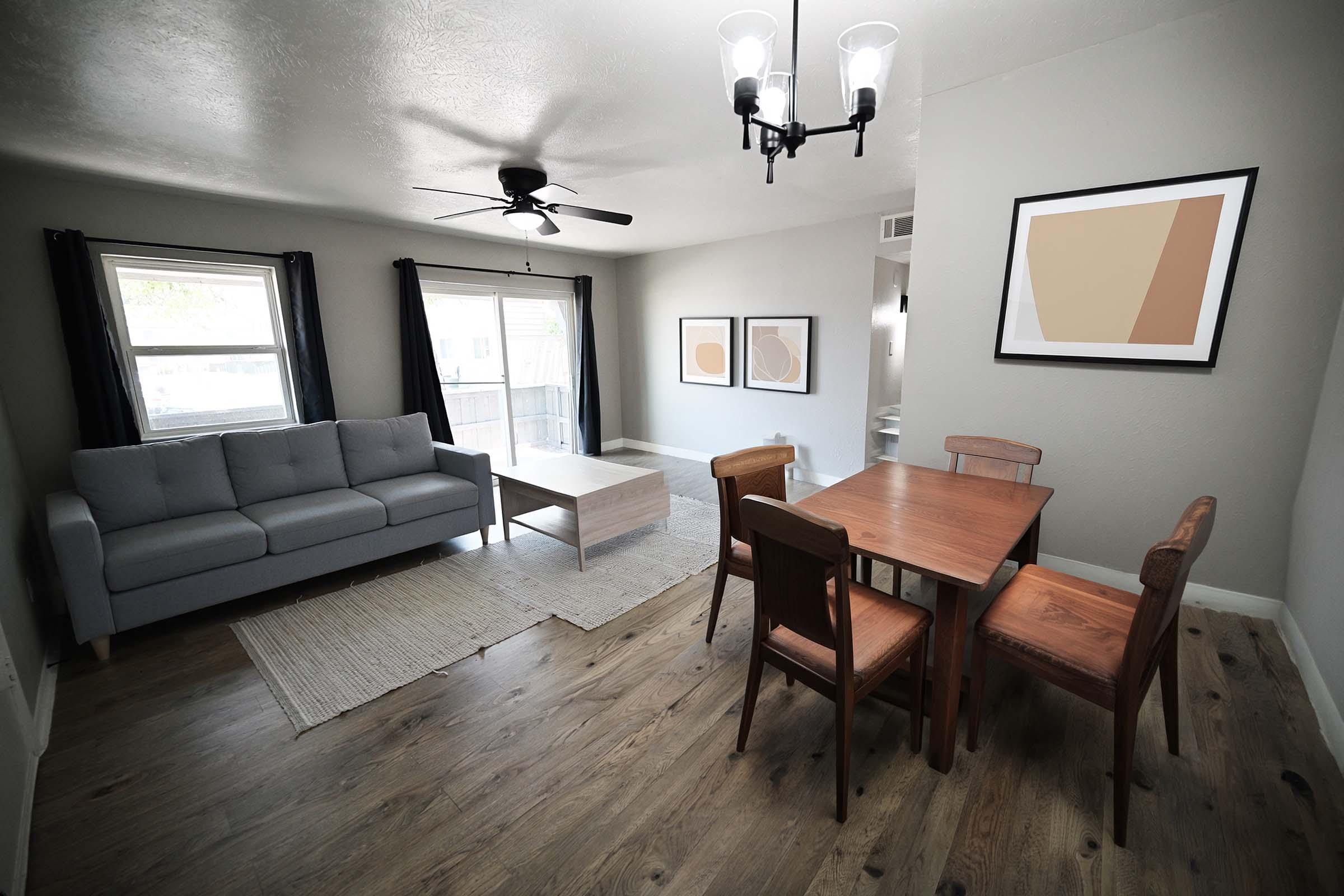 A modern living space featuring a gray sofa, a wooden coffee table, and a dining table with four chairs. Large windows provide natural light, and the walls are painted in a soft gray tone. Two abstract art pieces frame the area, adding a contemporary feel to the room.