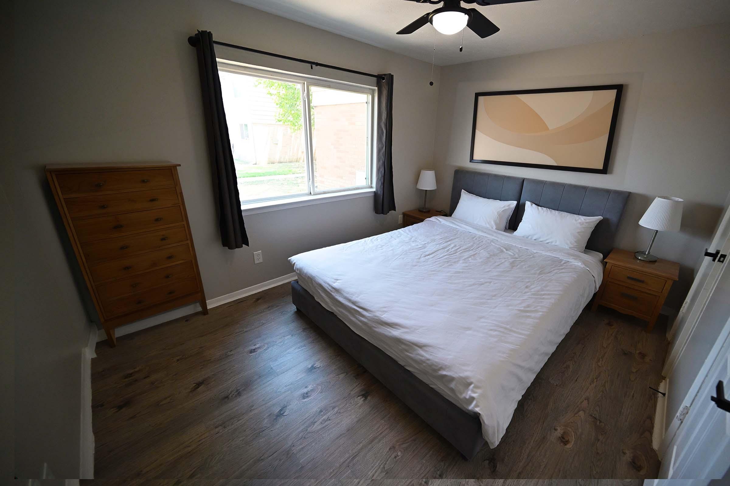 A cozy bedroom featuring a gray bed with white bedding, two nightstands with lamps on either side, a wooden dresser, and a large window letting in natural light. The walls are painted a soft gray, and there is a modern artwork piece above the bed, adding a touch of decor to the space.