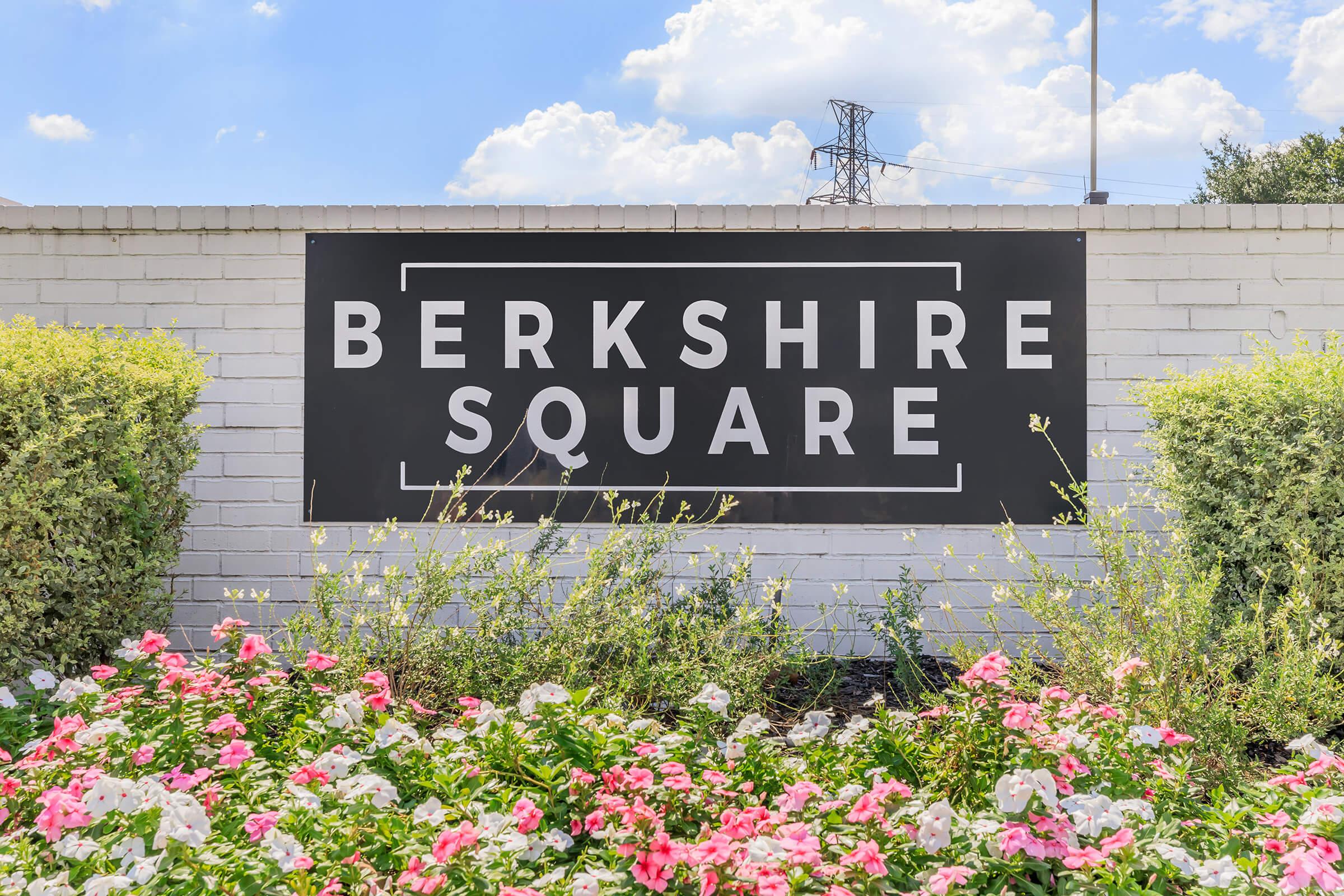 Signage for Berkshire Square, prominently displayed on a white wall, surrounded by colorful pink and white flowers. The background features a clear blue sky with fluffy clouds, along with power lines in the distance.