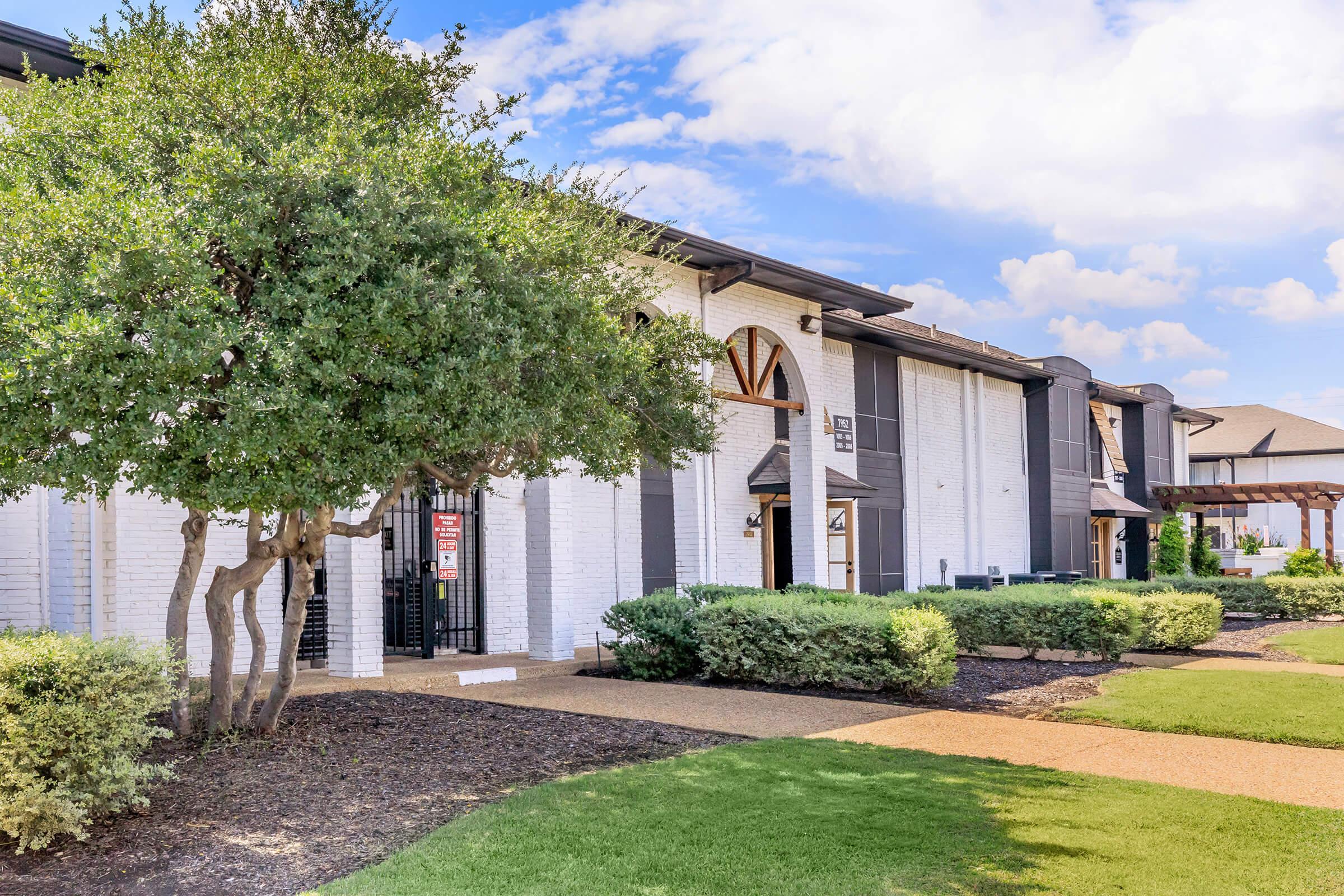 A picturesque view of a modern building surrounded by well-maintained landscaping, featuring a pathway lined with bushes and trees. The building has a white brick exterior with dark accents and large windows, set against a blue sky with scattered clouds.