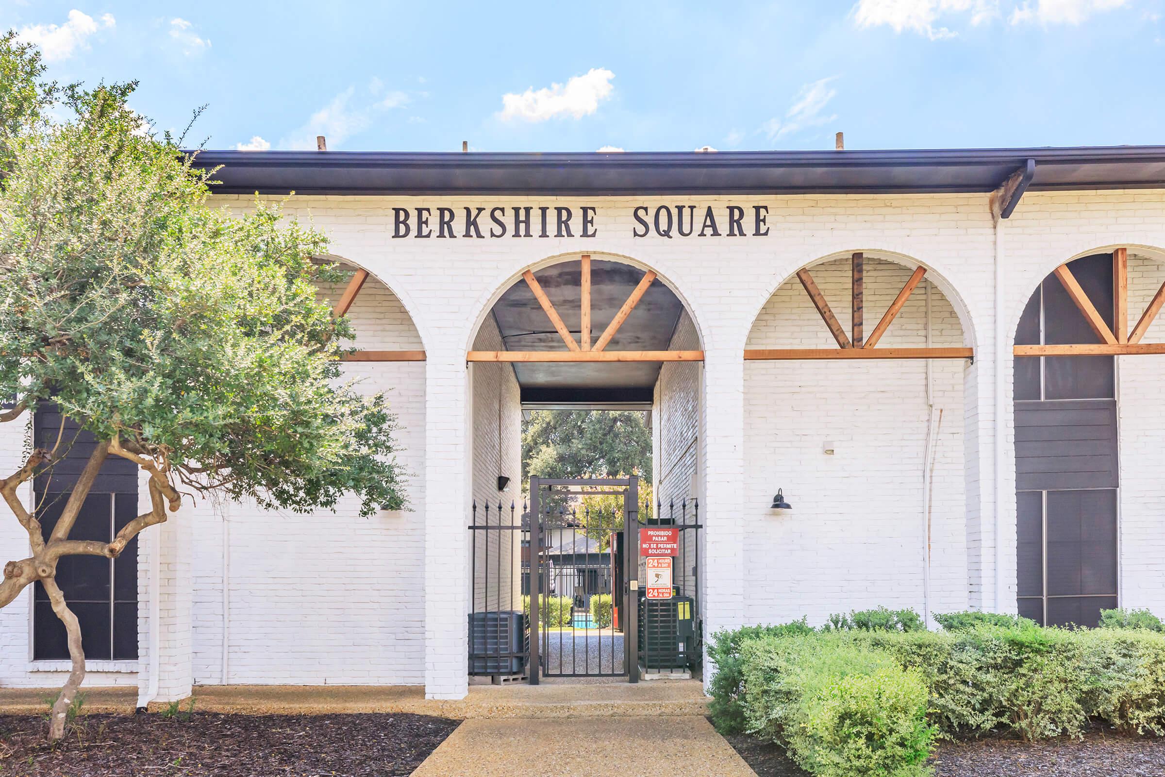 A white brick building with arched entrances, labeled "Berkshire Square" at the top. The entrance is fenced, and there are bushes and trees in the foreground. The sky above is blue with some clouds.