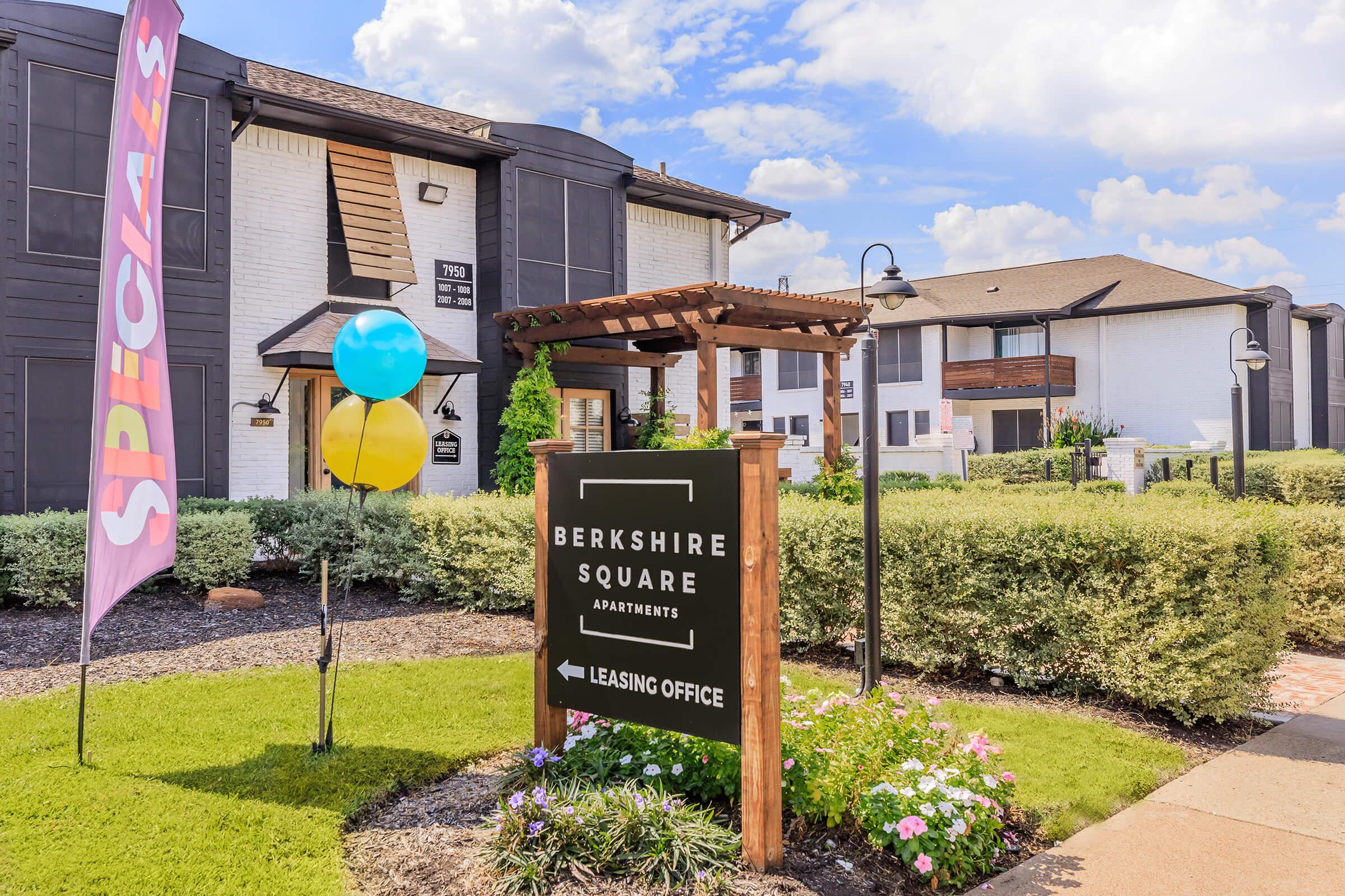 Sign for Berkshire Square Apartments with arrows pointing to the leasing office. Colorful balloons and a "Specials" banner enhance the outdoor area, which features well-maintained landscaping and a clear blue sky. The buildings are modern with a blend of dark and light exterior.