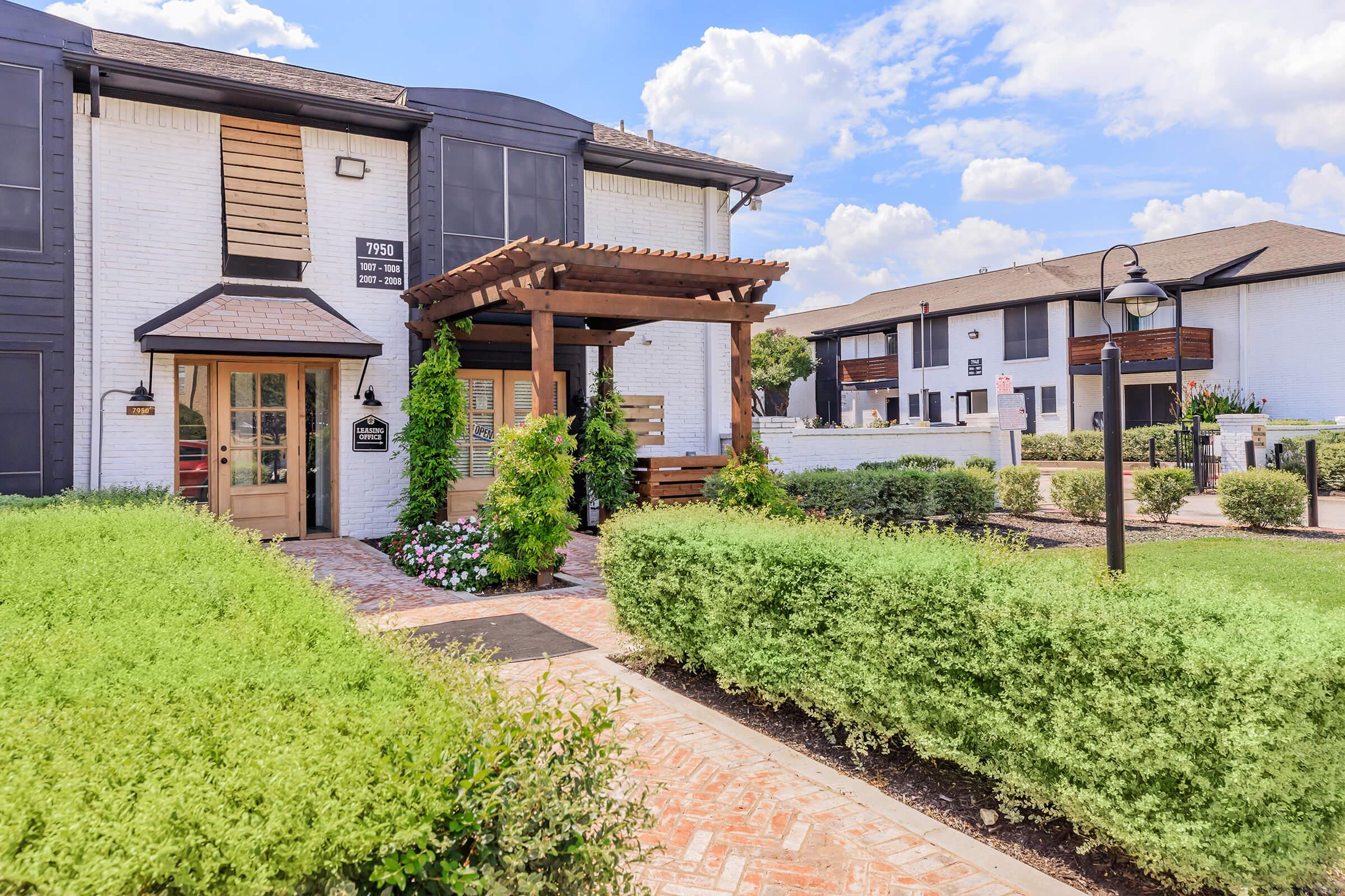 A quaint apartment entrance with a wooden pergola surrounded by lush greenery and colorful flowers. A brick walkway leads to the door, and a street lamp stands nearby. In the background, multiple apartment buildings can be seen under a bright blue sky with fluffy white clouds.