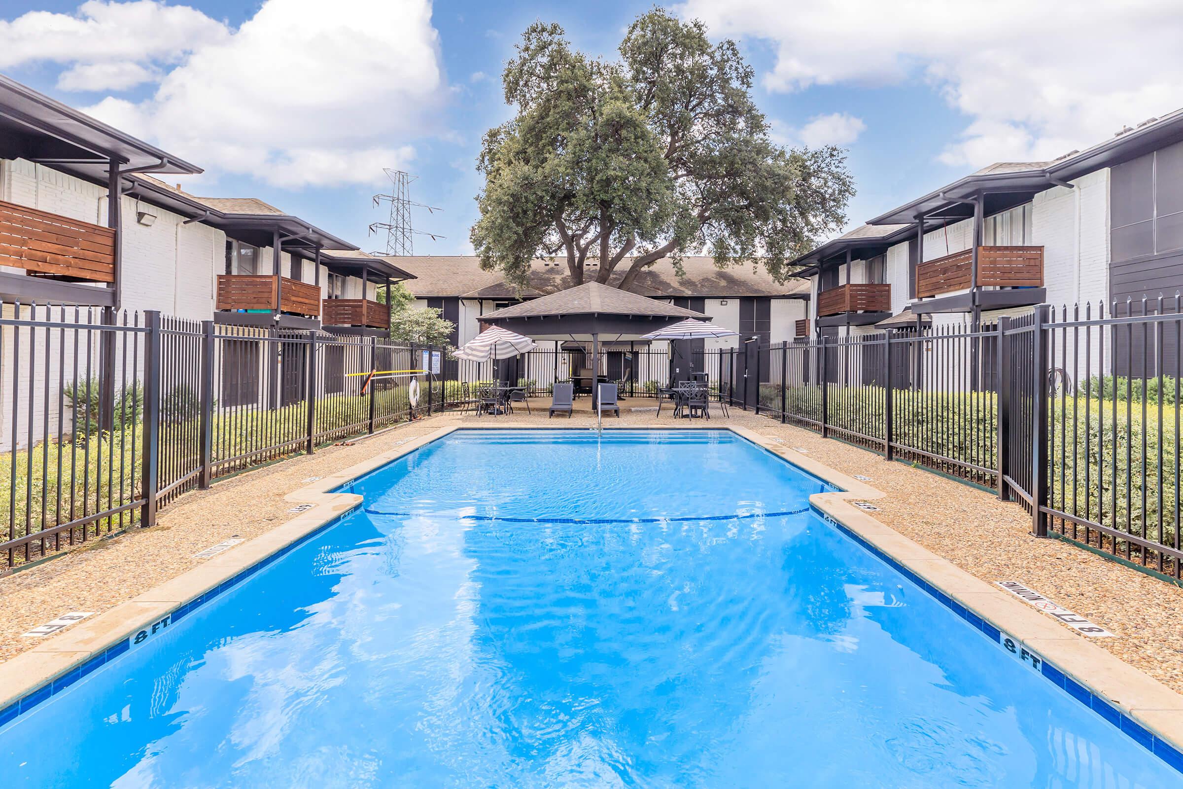 A clear blue swimming pool surrounded by a fenced area, with sun loungers and an umbrella for shade. In the background, there are white apartment buildings with balconies and green landscaping, under a partly cloudy sky.