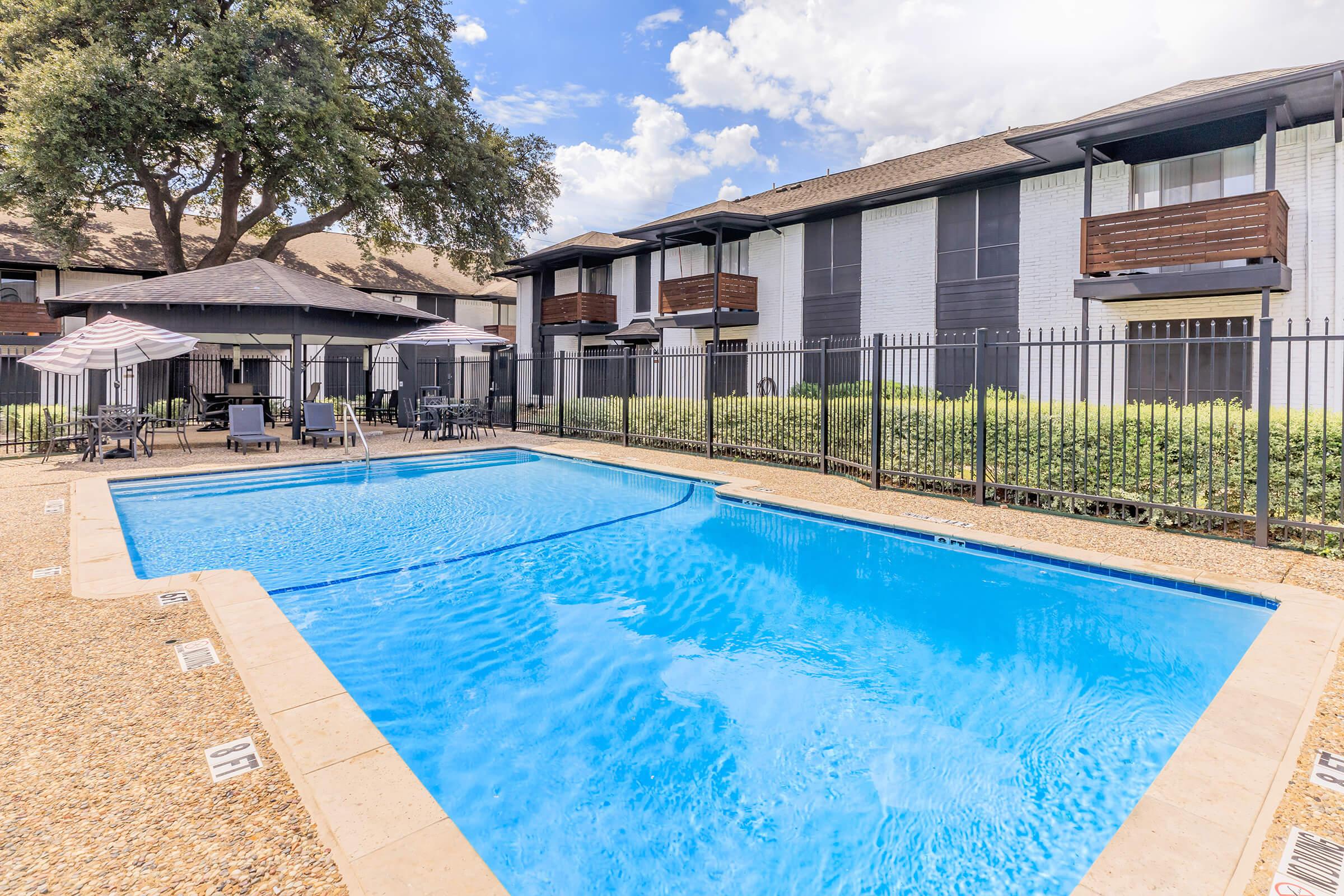 A clear blue swimming pool surrounded by a fenced area, with several lounge chairs and umbrellas nearby. In the background, there are modern apartment buildings with balconies, and a large tree providing shade. The sky is partly cloudy, creating a bright and inviting atmosphere.