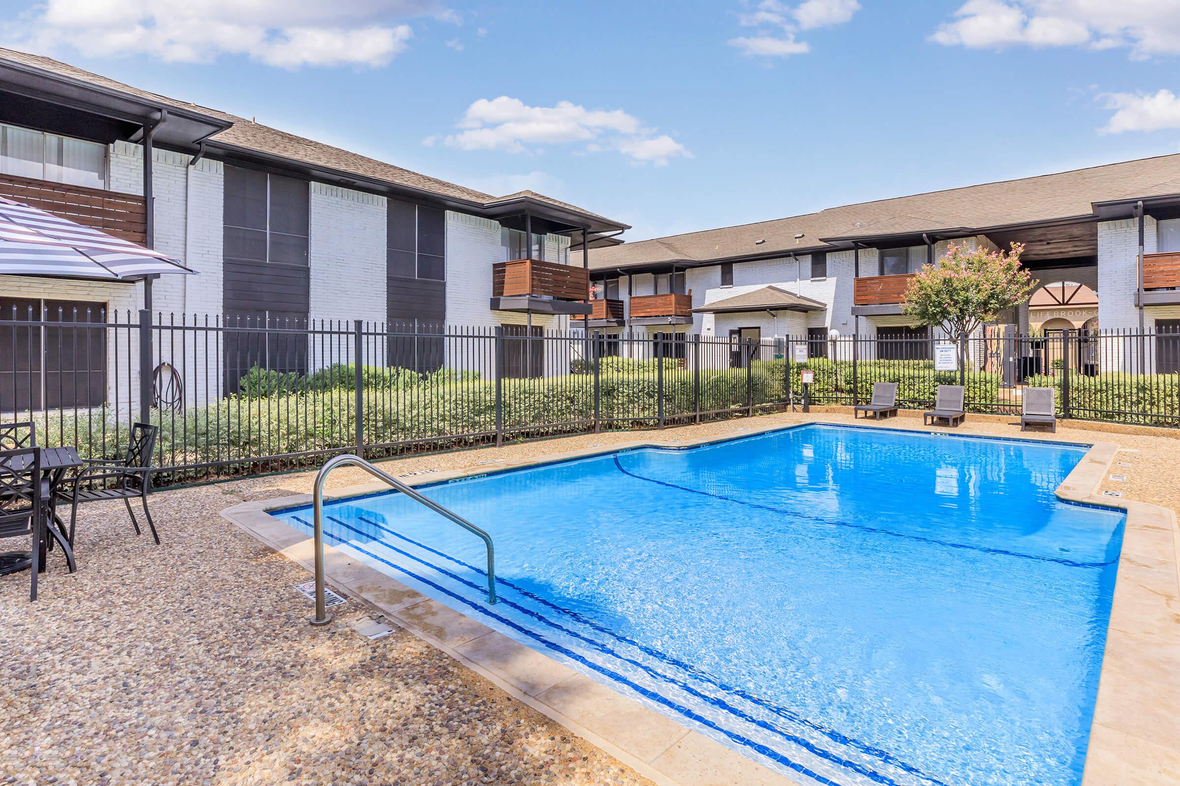 A clear blue swimming pool surrounded by a fenced area. Sun loungers are placed around the pool, and there are leafy shrubs in the background. The setting includes two-story apartment buildings with a modern design and balconies, under a bright blue sky with fluffy white clouds.