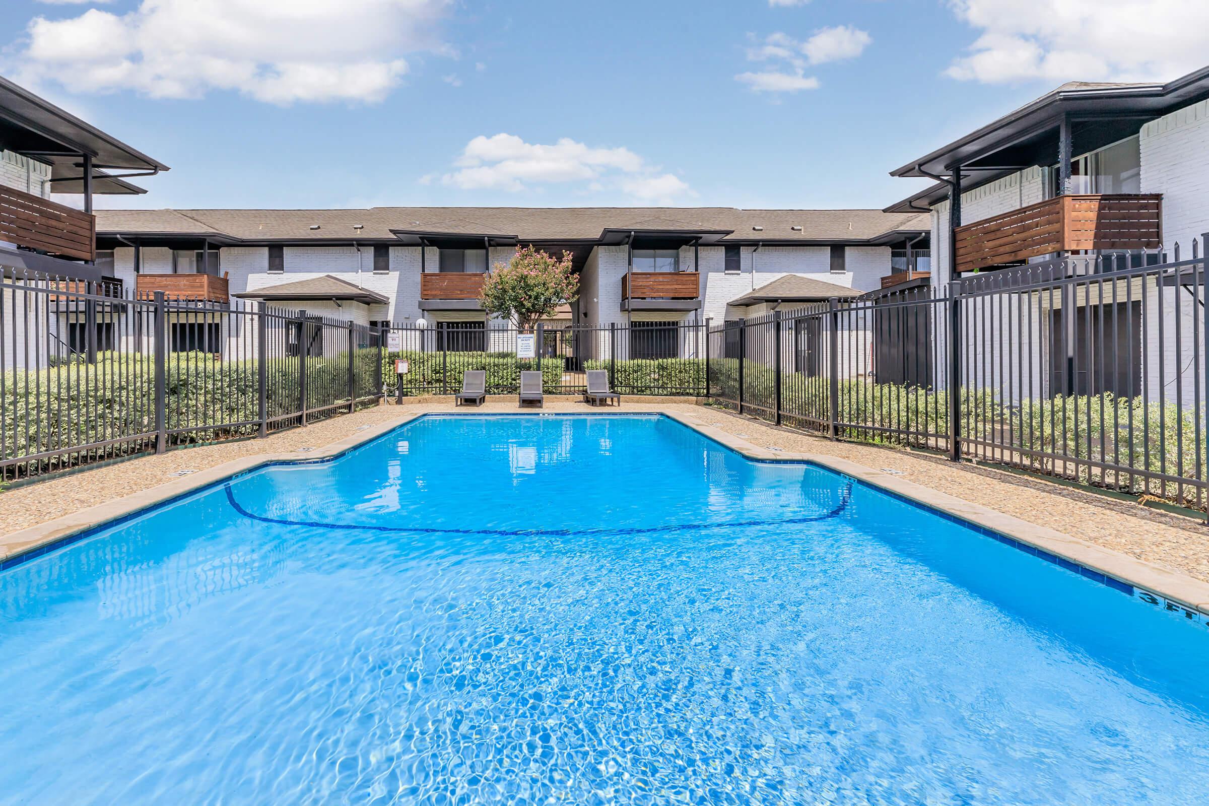 A clear blue swimming pool surrounded by a black fence, with lounge chairs on one side. In the background, there are two-story apartment buildings with balconies, and lush greenery lining the edges of the pool area. The sky is bright with a few fluffy clouds.