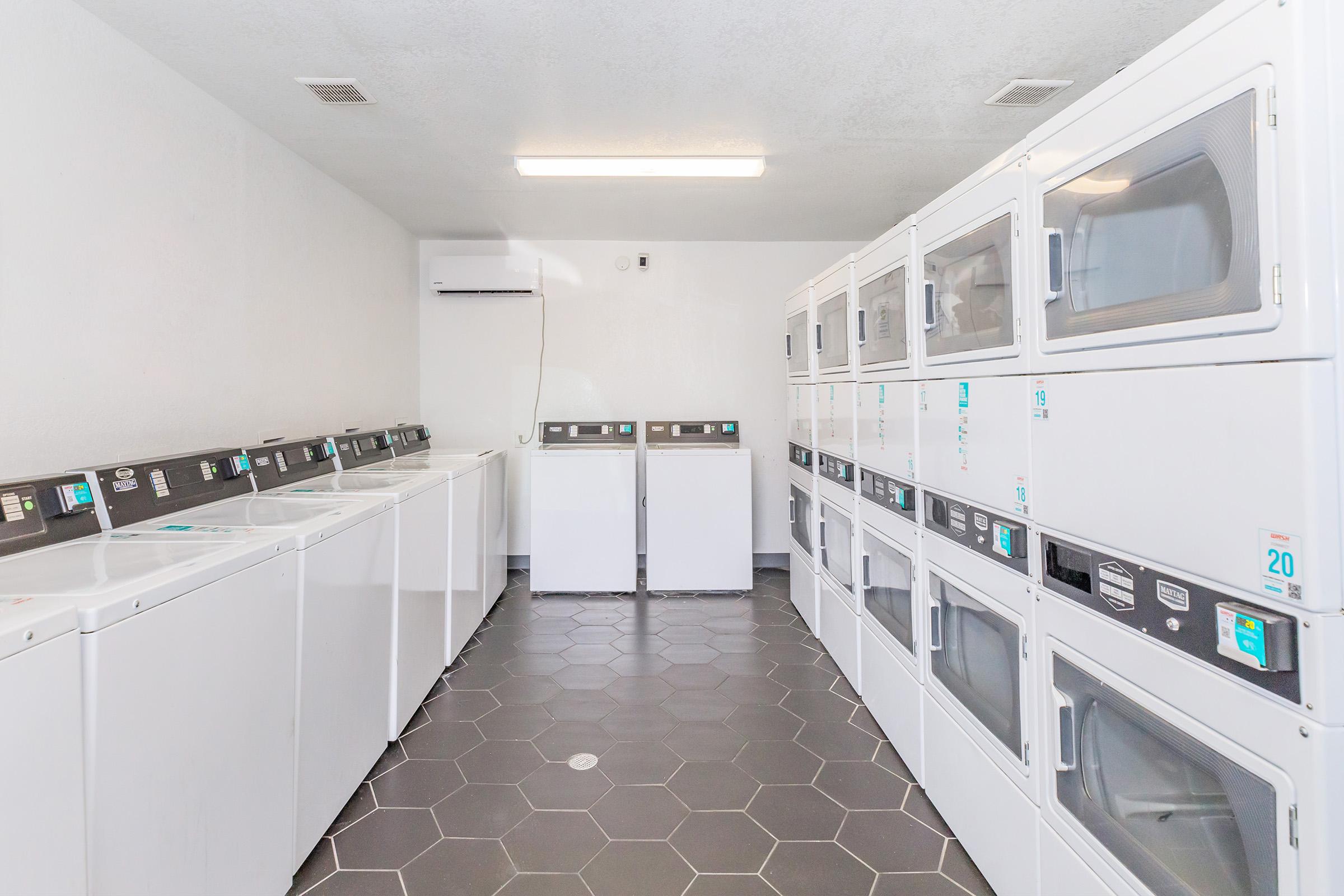 A clean and well-lit laundry room featuring multiple washing machines and dryers lined up against the wall. The floor is covered with hexagonal tiles, and there are two rows of machines with digital controls. The room is simple and functional, designed for residential laundry use.