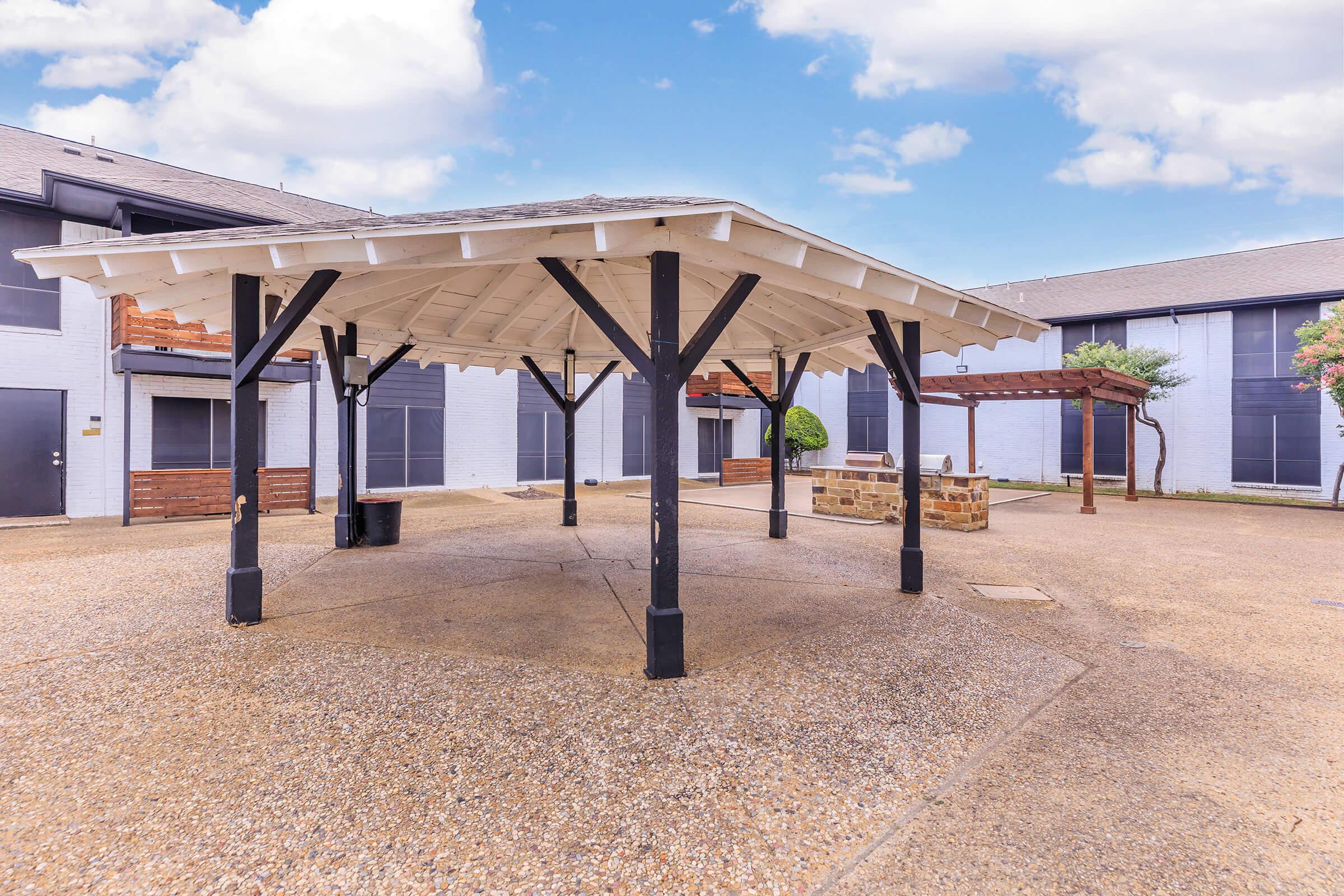 A covered outdoor gazebo with a white wooden frame and a gravel floor, surrounded by buildings. There are benches and planters nearby, and a clear blue sky with fluffy white clouds overhead, creating a serene courtyard atmosphere.