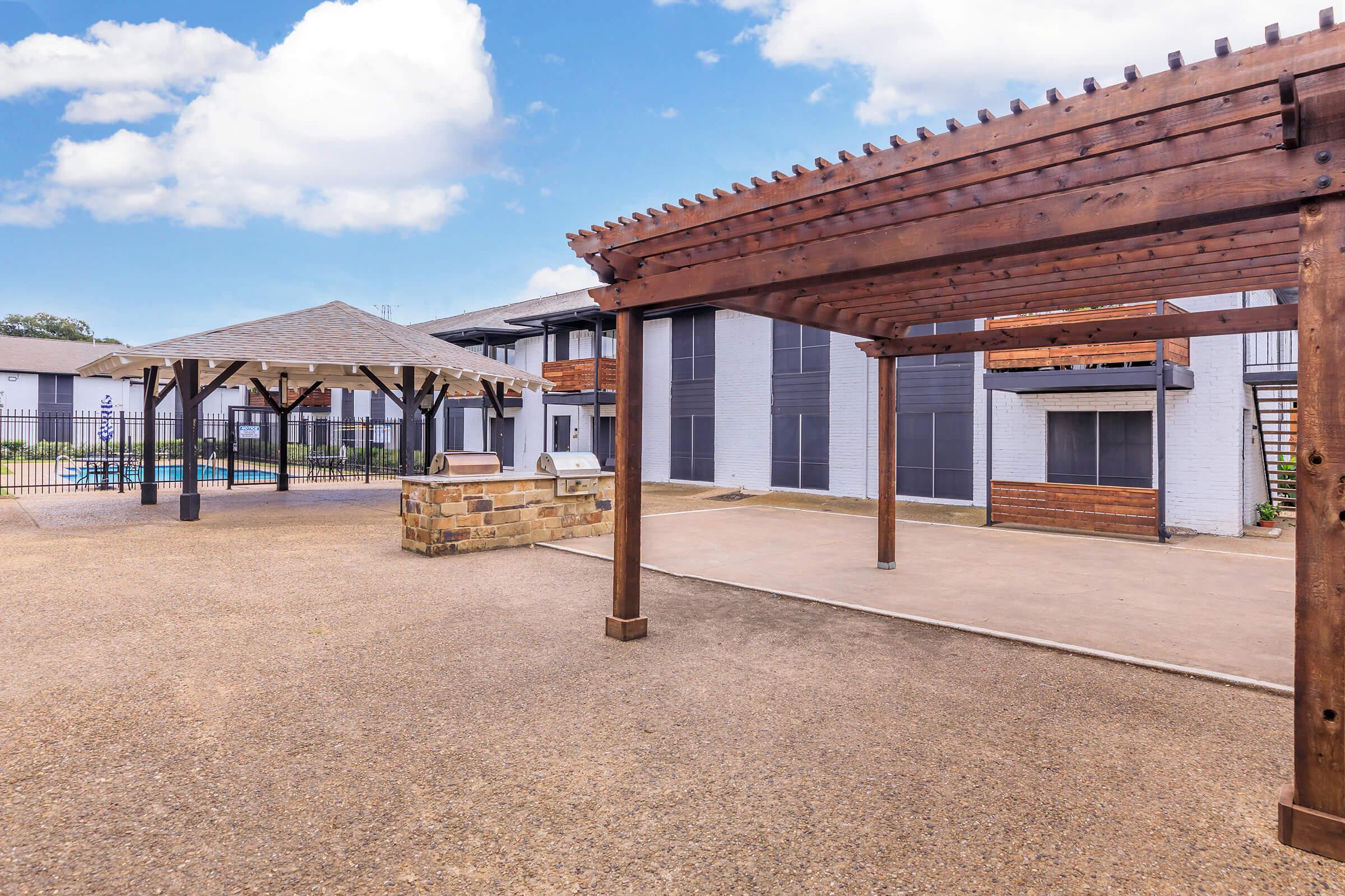 A view of a courtyard area featuring a wooden pergola, a gazebo, and a swimming pool surrounded by a fence. The background shows a multi-story building with balconies. The sky is partly cloudy, providing a bright atmosphere. The ground is paved with gravel, creating a warm and inviting outdoor space.