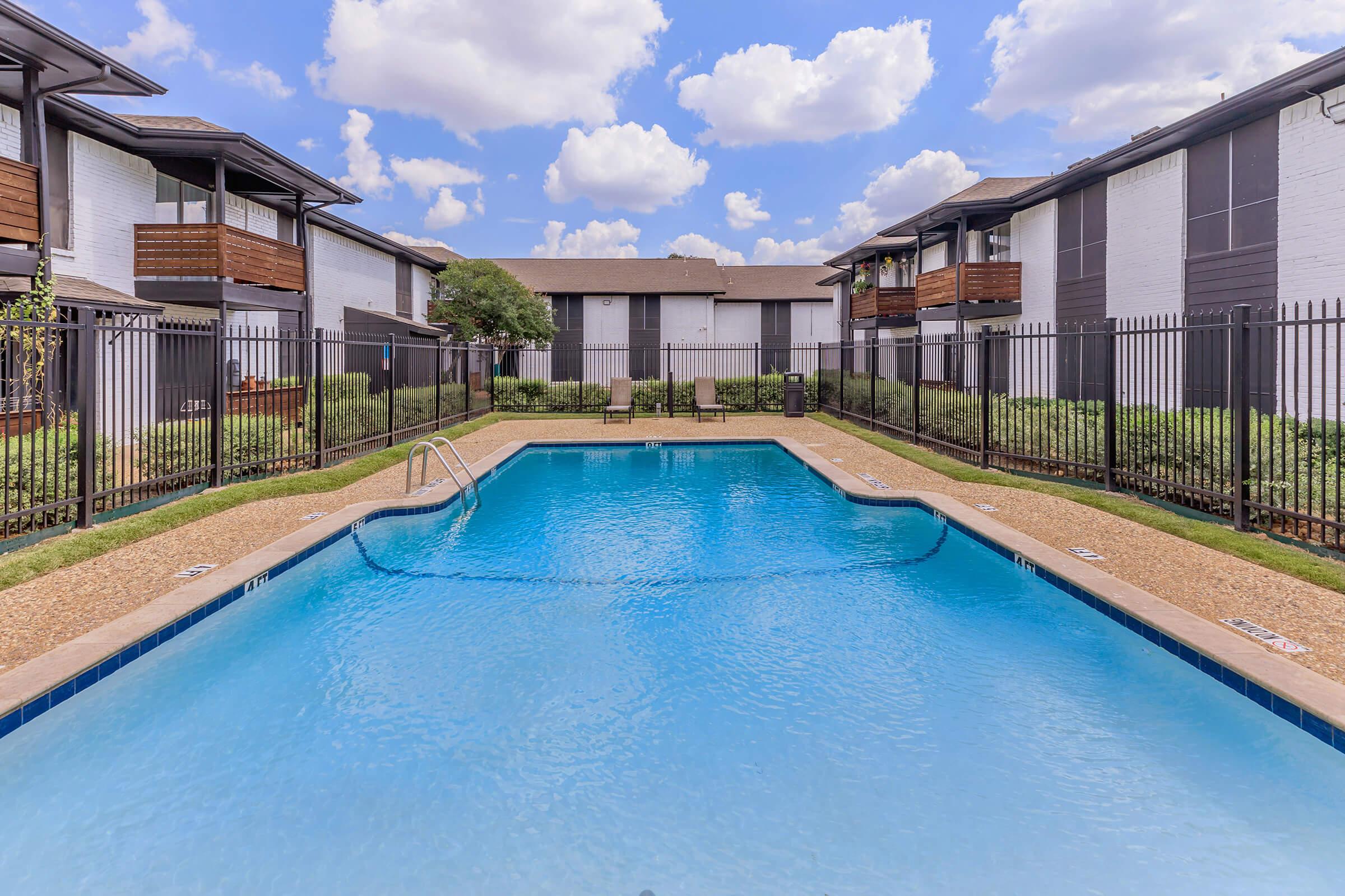 A sunny outdoor swimming pool surrounded by green grass and shrubs, located in a residential complex. The pool has a clear blue water and a ladder for entry, with two lounge chairs nearby. In the background, there are two-story apartment buildings with balconies under a partly cloudy sky.