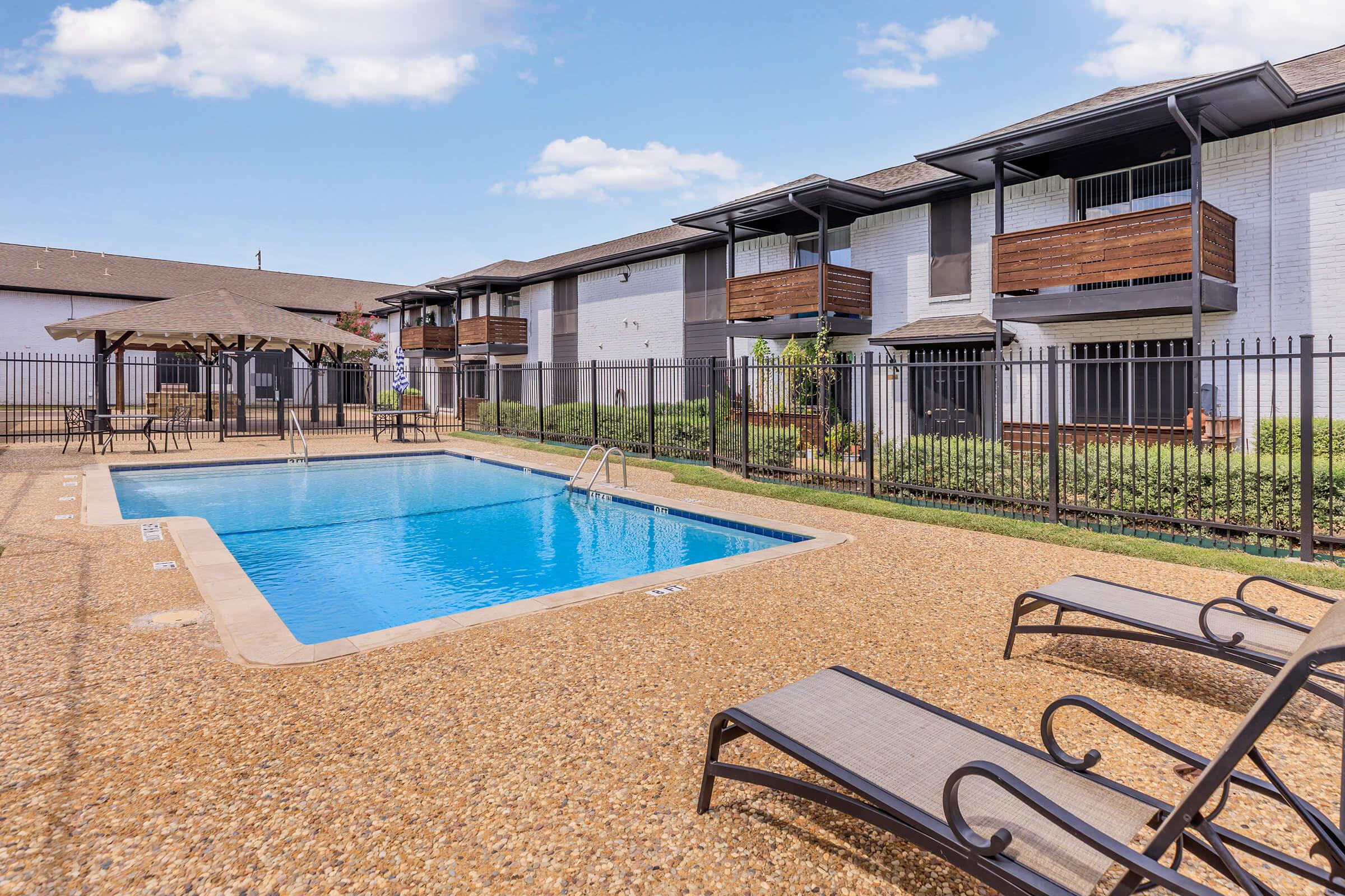 A clear blue swimming pool surrounded by a fenced area, with two lounge chairs on the side. In the background, there are two apartment buildings with wooden balconies. The scene is sunny, showcasing well-maintained landscaping and a serene atmosphere.