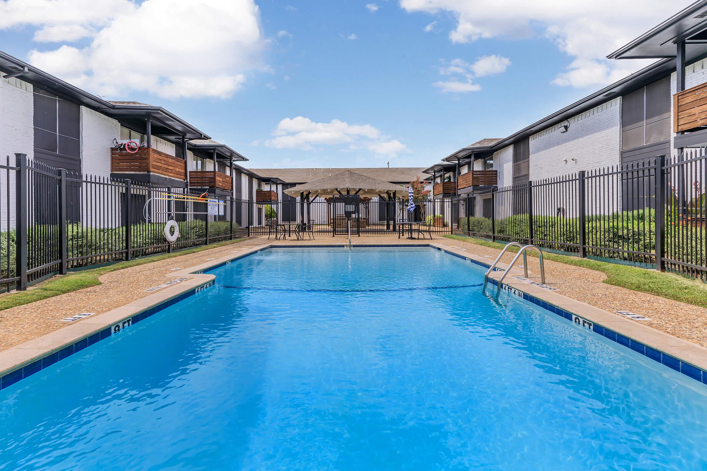 A view of a clean swimming pool surrounded by modern apartment buildings. The pool has a ladder, lounge chairs nearby, and is framed by a black fence. Lush greenery is present along the edges, with a clear blue sky and a few clouds above, creating a serene atmosphere.