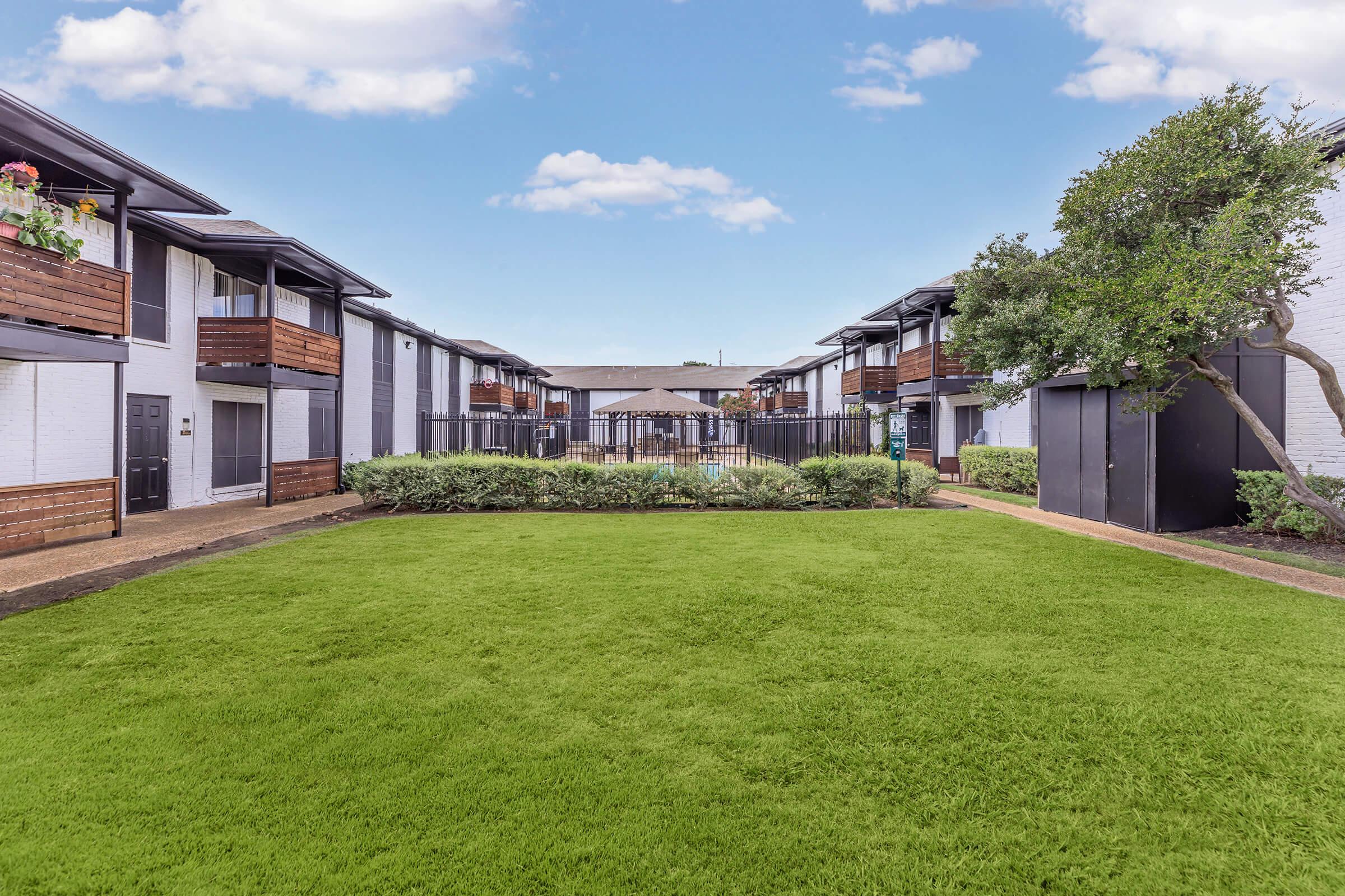 A landscaped courtyard surrounded by residential buildings. The area features well-maintained green grass, colorful flower boxes on the balconies, and a gated entrance visible in the background under a clear blue sky.