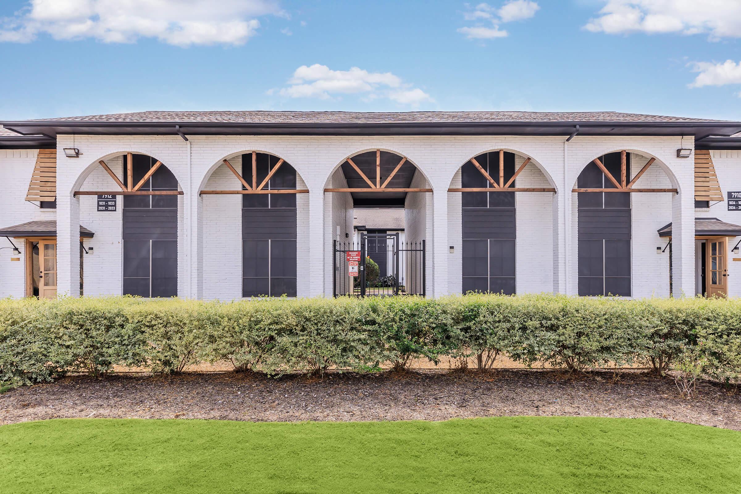 A modern building with a white exterior featuring large arched windows and wooden accents. The entrance is gated, flanked by greenery and a manicured lawn. The sky above is clear and blue, adding to the bright ambiance of the scene.