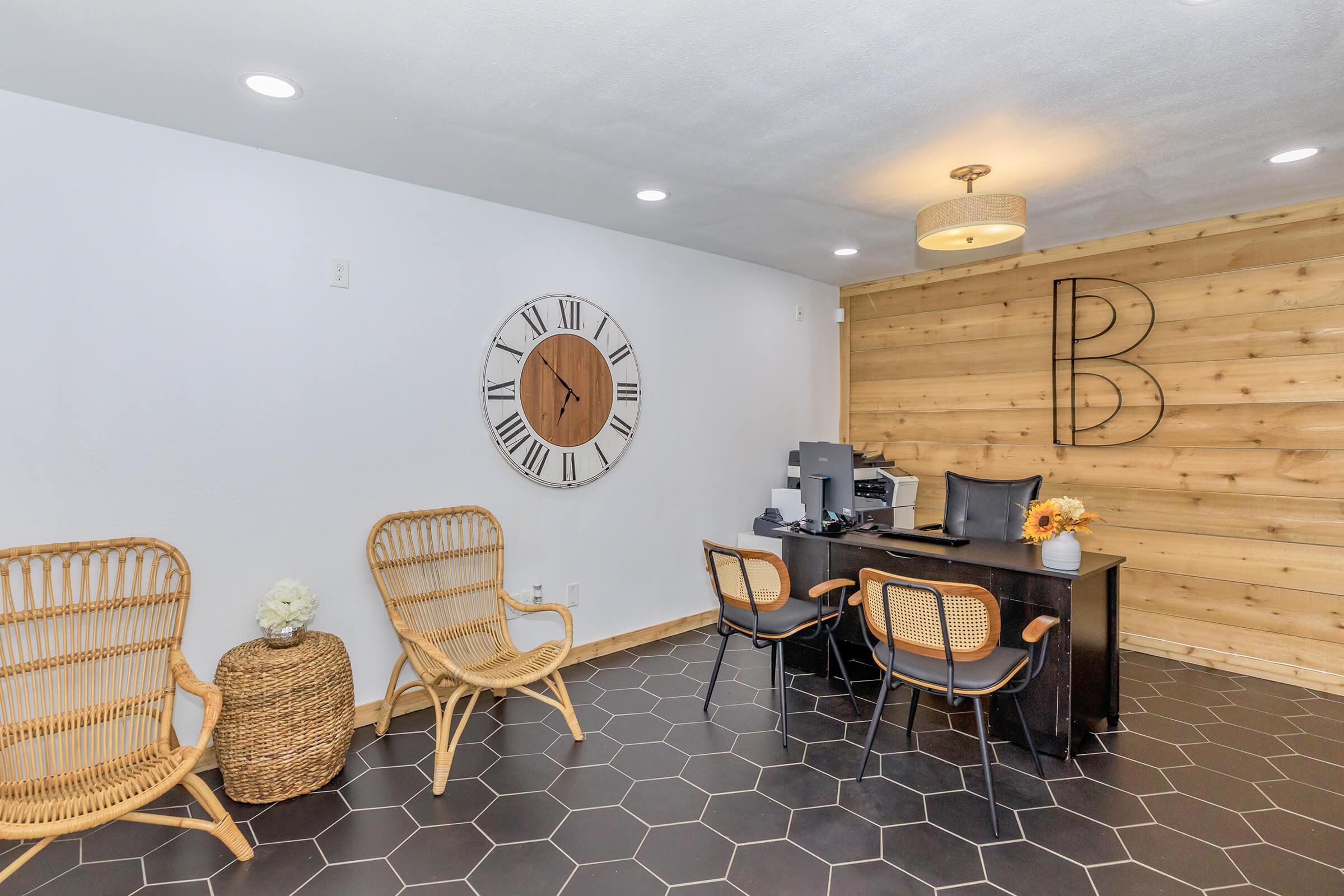 A modern reception area featuring a wooden accent wall with a large letter "B," a round clock, a desk with a black chair and computer, and two rattan chairs. The floor is tiled in a hexagonal pattern, and there is a decorative basket and a flower arrangement on display.