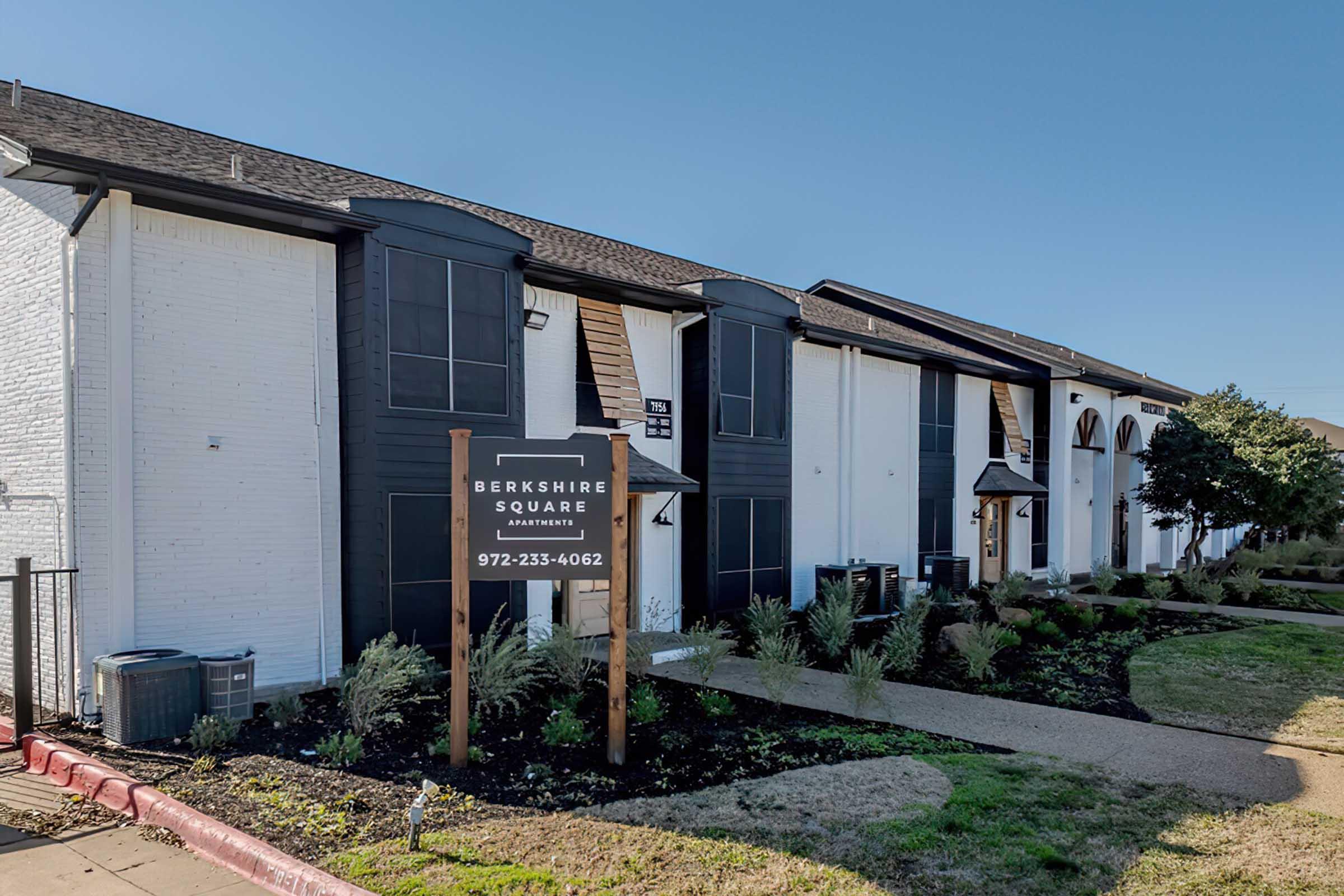 A view of Berkshire Square Apartments, showcasing a row of modern apartment buildings with a prominent sign displaying the name and contact number. The area is landscaped with plants and grass, and there's a clear blue sky in the background.