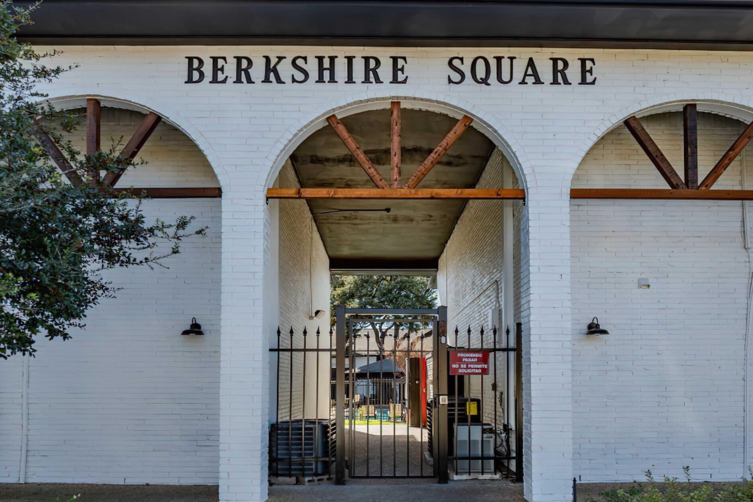 Entrance to Berkshire Square featuring a white brick facade and arched structures. The area is gated, and a red sign is visible near the entrance. In the background, there is a glimpse of a pool area, surrounded by landscaped greenery.