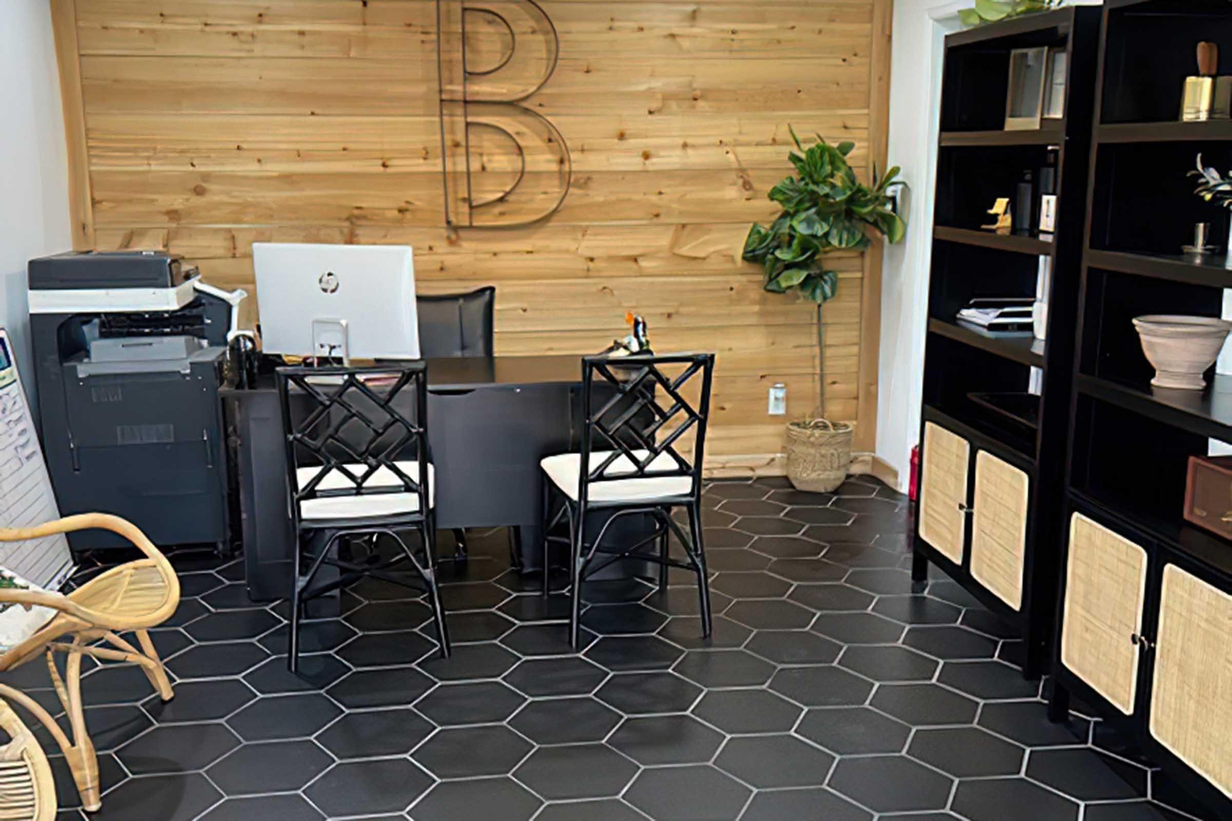 A modern office space featuring a wooden accent wall with the letter "B," a sleek black desk with two black and white chairs, a printer, a potted plant, and a bookshelf with various decorative items. The floor is designed with hexagonal tiles, creating a contemporary atmosphere.
