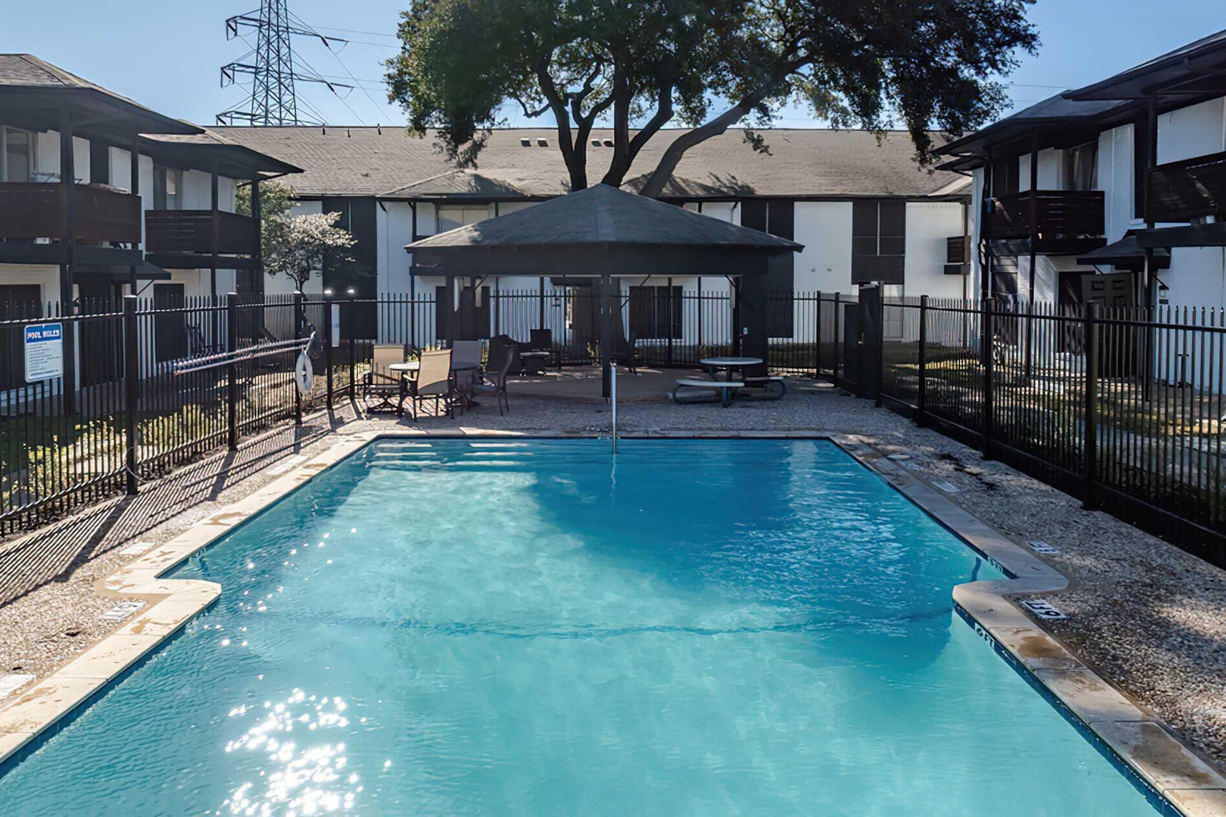 A clear swimming pool surrounded by a black iron fence, with lounge chairs and a shaded gazebo nearby. The scene features a well-maintained courtyard of an apartment complex, with trees and a utility pole in the background, under a bright blue sky.