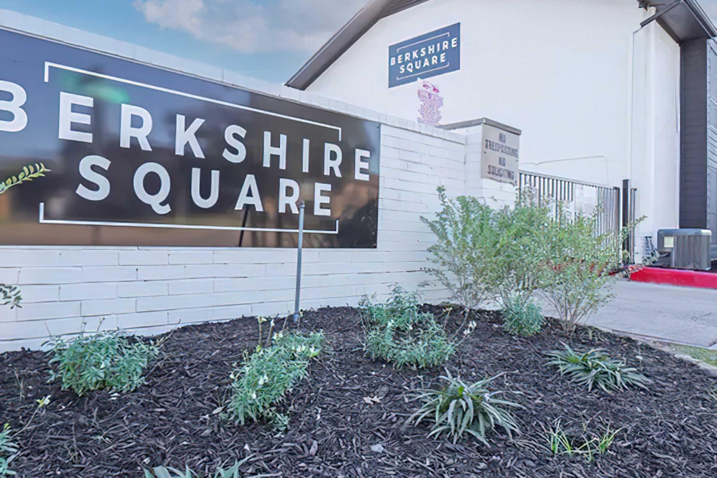 Signage for Berkshire Square in front of a landscaped area with green plants and mulch, accompanied by a white building in the background. The environment appears well-maintained and welcoming.