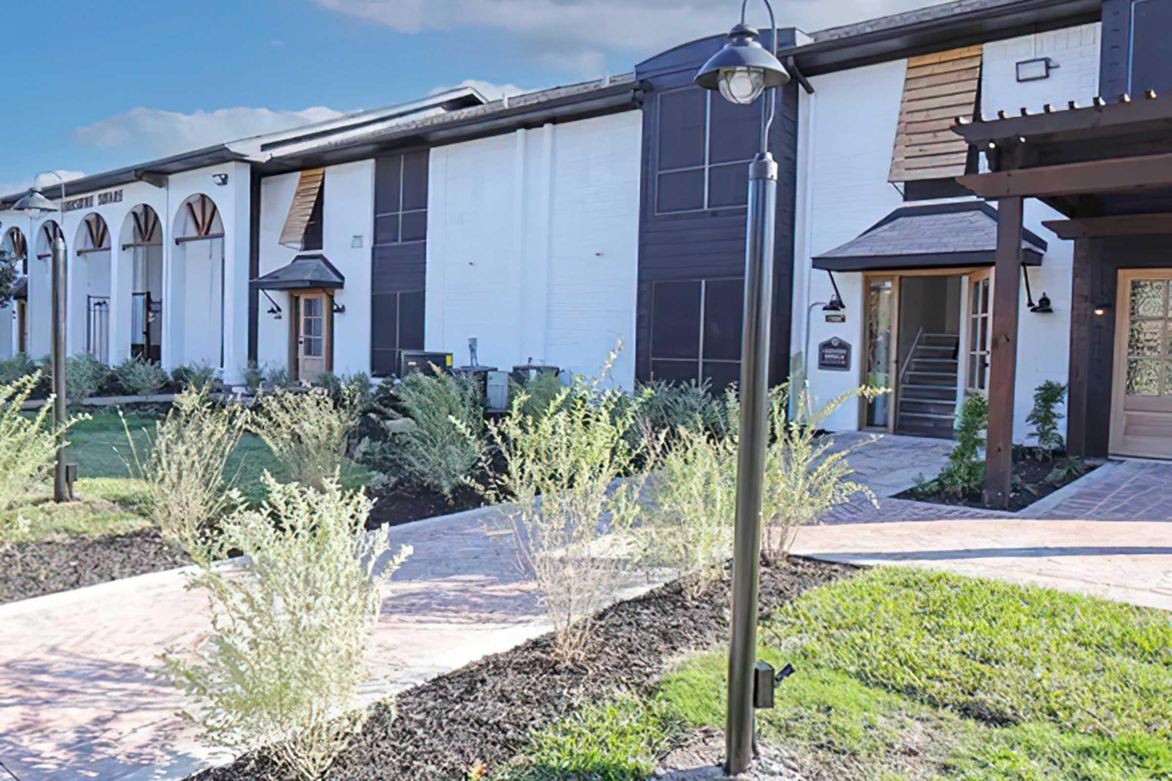 A modern building with a combination of white and dark exterior features. There are pathways lined with plants, a lamp post, and an inviting entrance with large windows and a wooden awning. The landscaping is simple and well-maintained, creating a welcoming atmosphere.